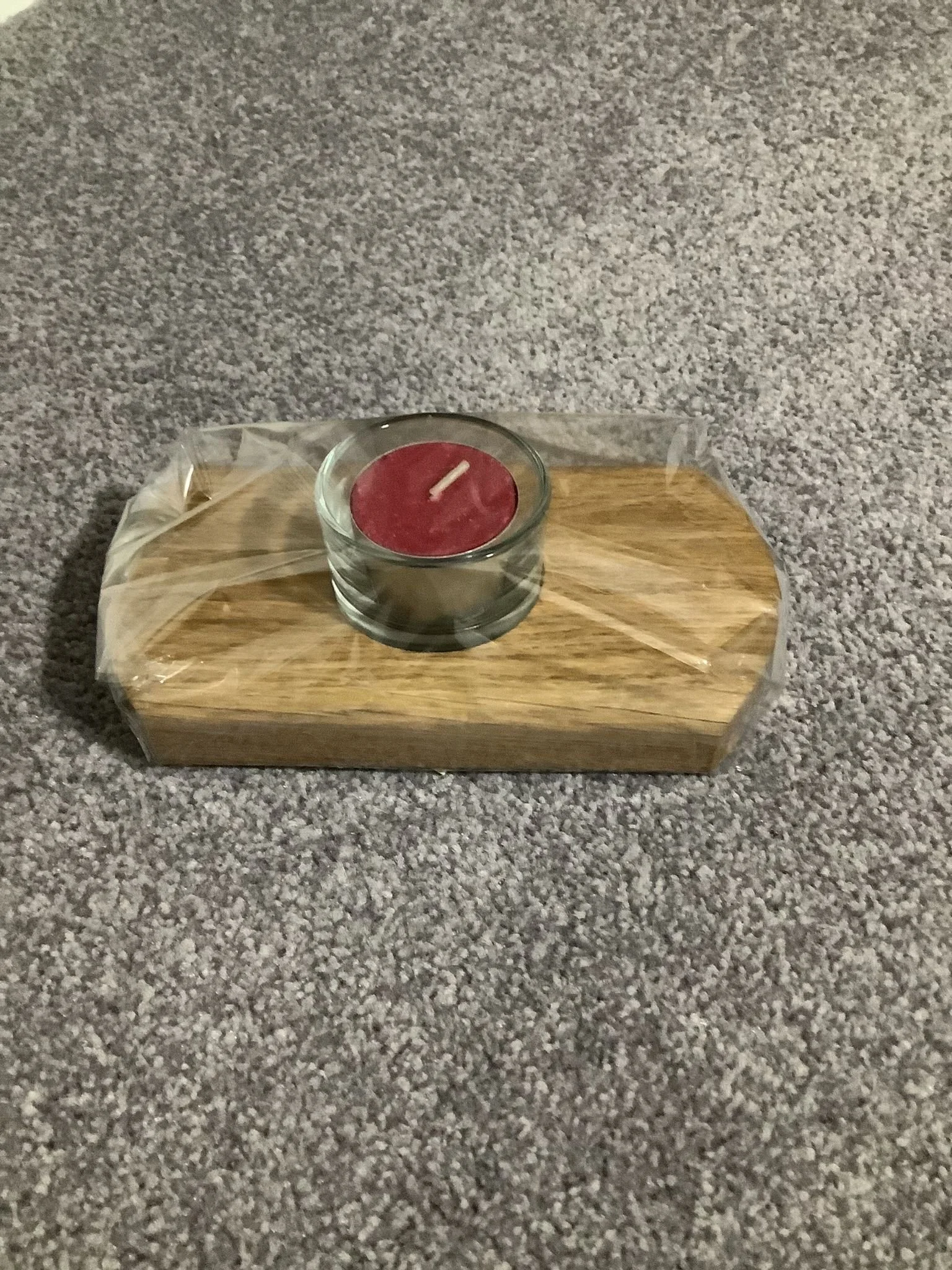Red candle in a glass holder on a wooden tray, wrapped in clear plastic, on a carpeted floor.