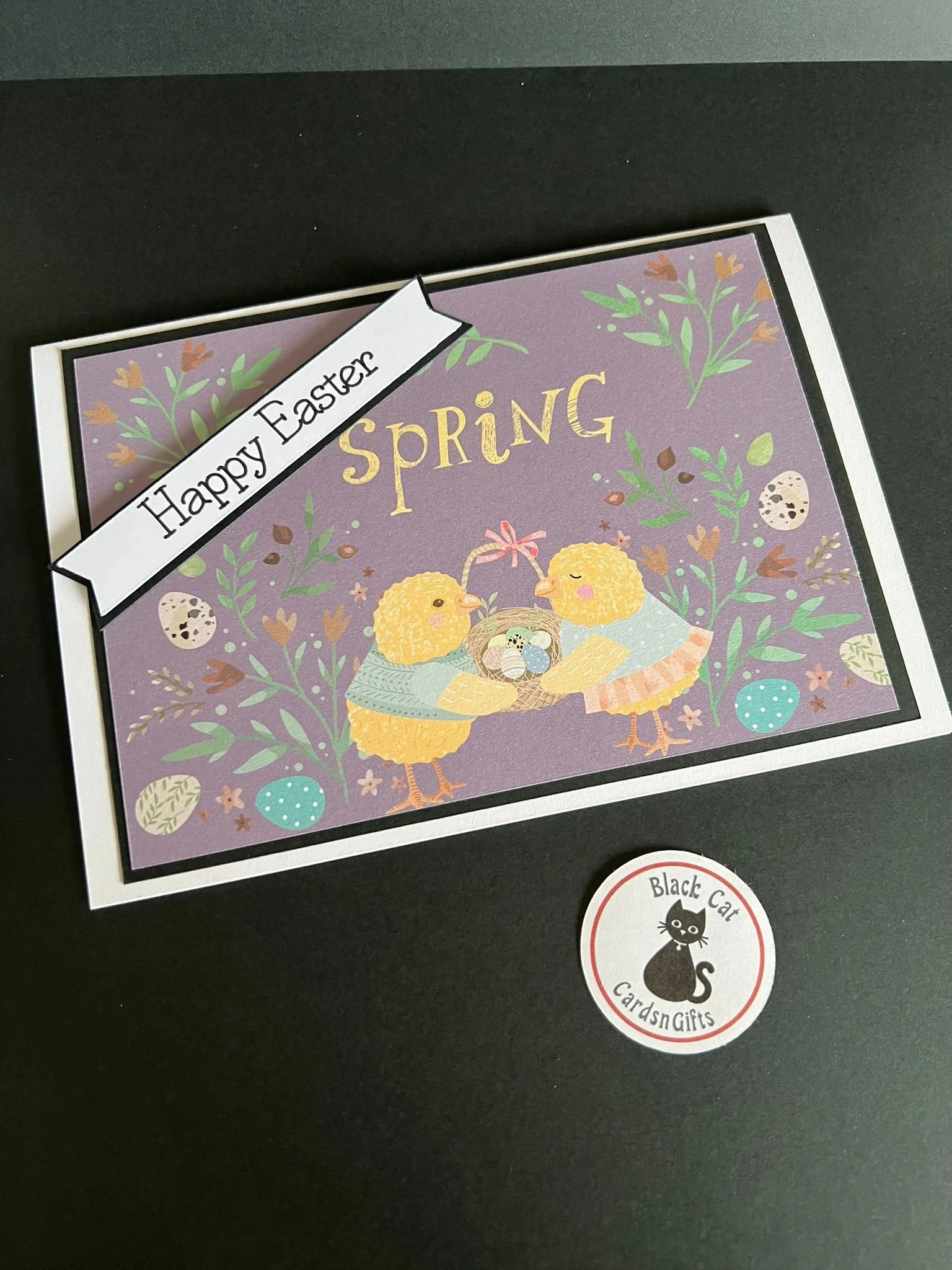 Decorative Easter greeting card with purple background, featuring two yellow chicks holding a nest with decorated eggs, surrounded by floral and egg illustrations, with a banner reading 'Happy Easter' and the word 'Spring' in gold letters. The card i