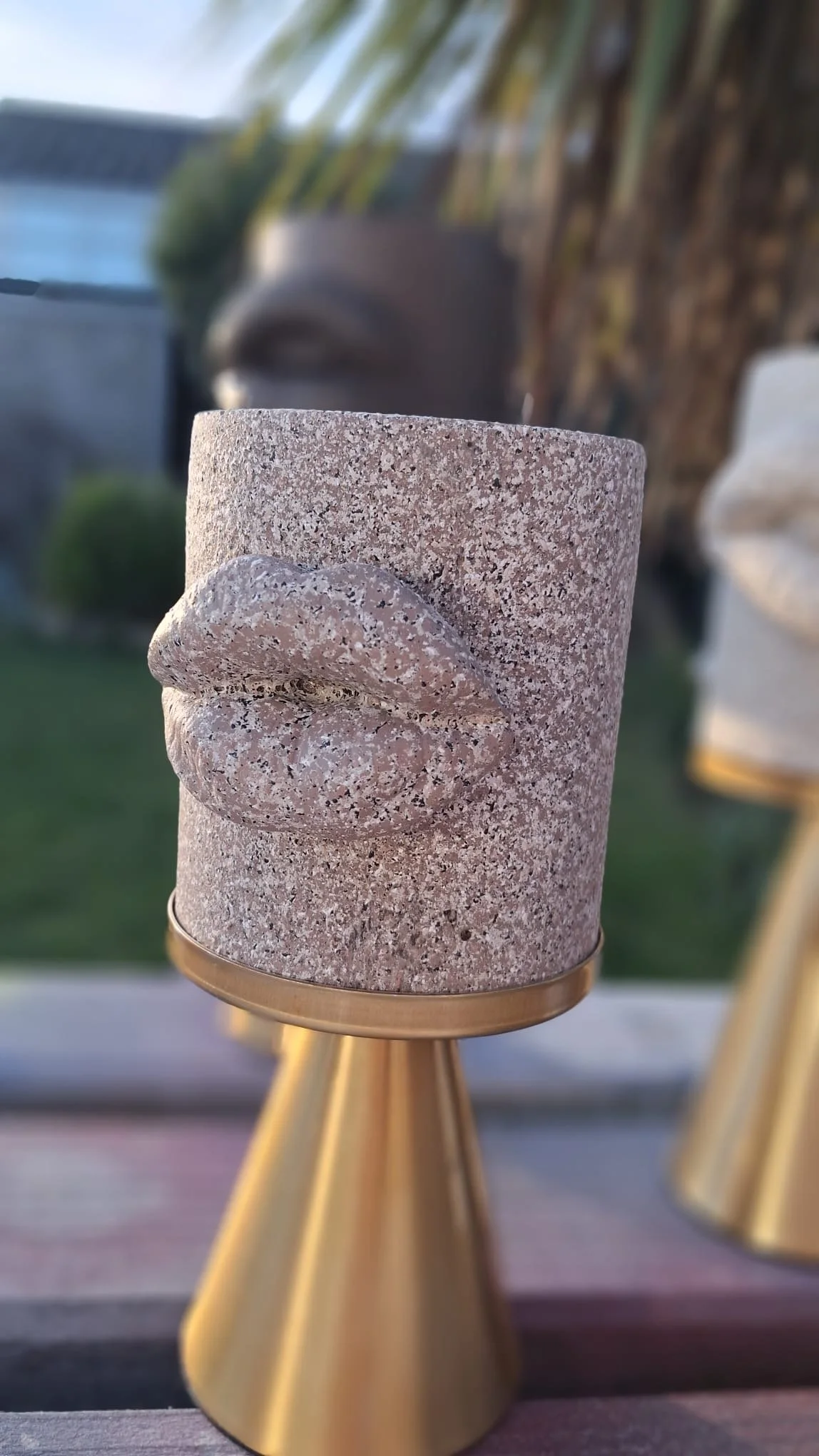 A concrete candle holder with a carved lips design on the front, positioned on a metallic stand outdoors, with a blurred garden and house background.