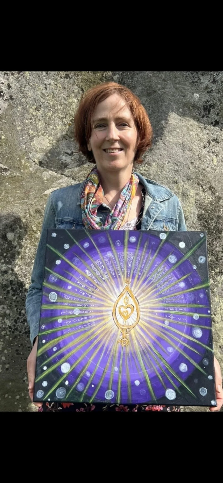 A woman with short red hair smiling, wearing a denim jacket and colorful scarf, holding a purple and gold painted canvas with a radiant design and symbolic symbols, standing in front of a rocky background.