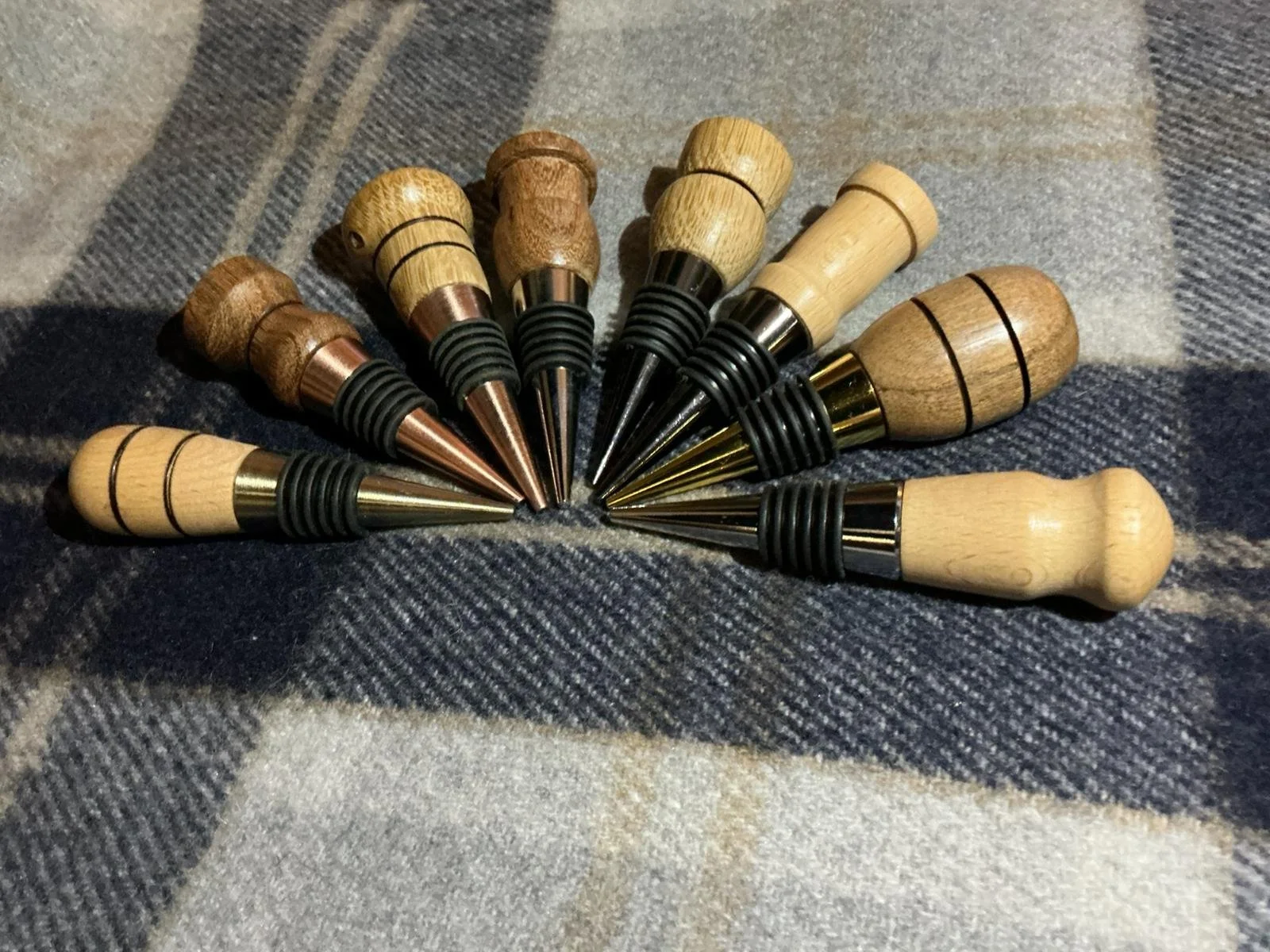 Set of ten bottle stoppers with wooden handles and metal tips, arranged in a semi-circle on a plaid fabric surface.