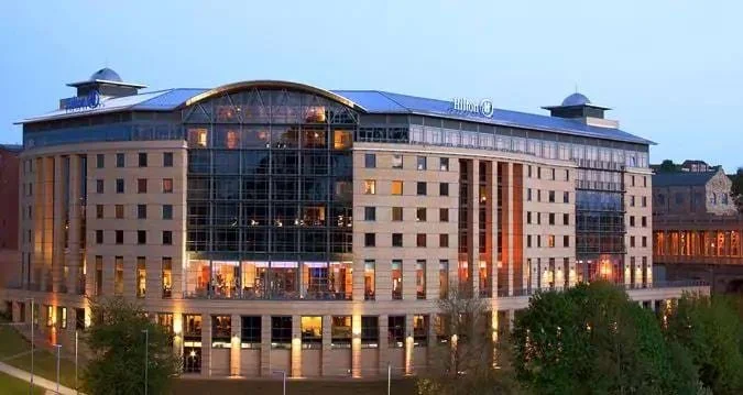 Exterior view of a modern Hilton hotel building during sunset with illuminated windows and greenery in the foreground.