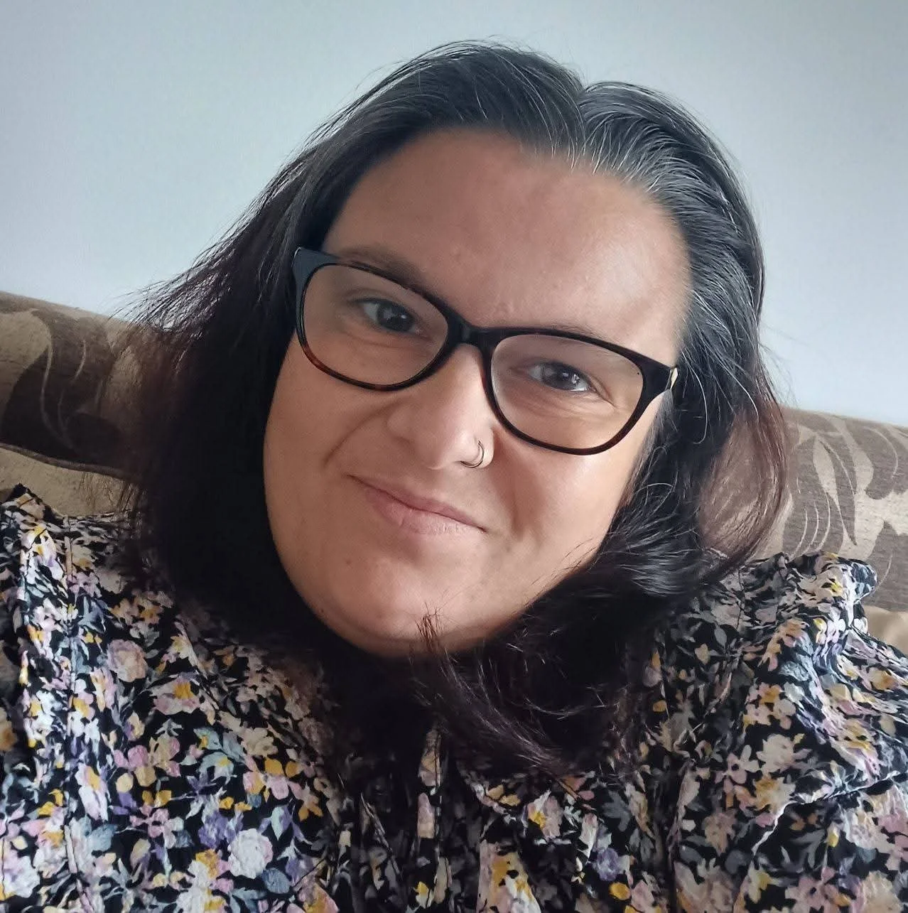 A person with shoulder-length dark hair, glasses, a nose ring, wearing a floral shirt, sitting on a patterned couch with a light-colored wall in the background.