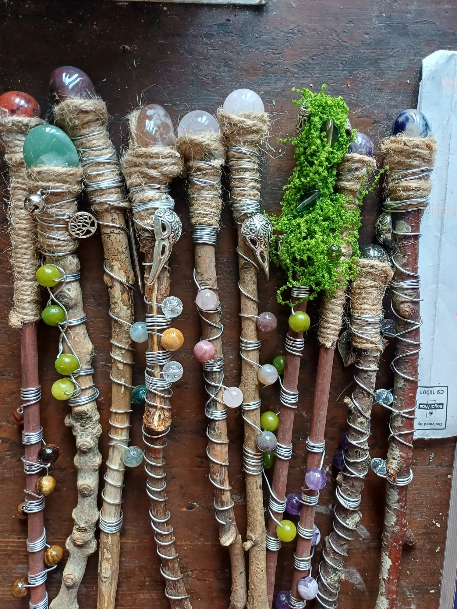 Collection of decorated sticks wrapped with wire and adorned with colorful beads and metal charms, with some green moss or foliage on one of the sticks, laid on a wooden surface.