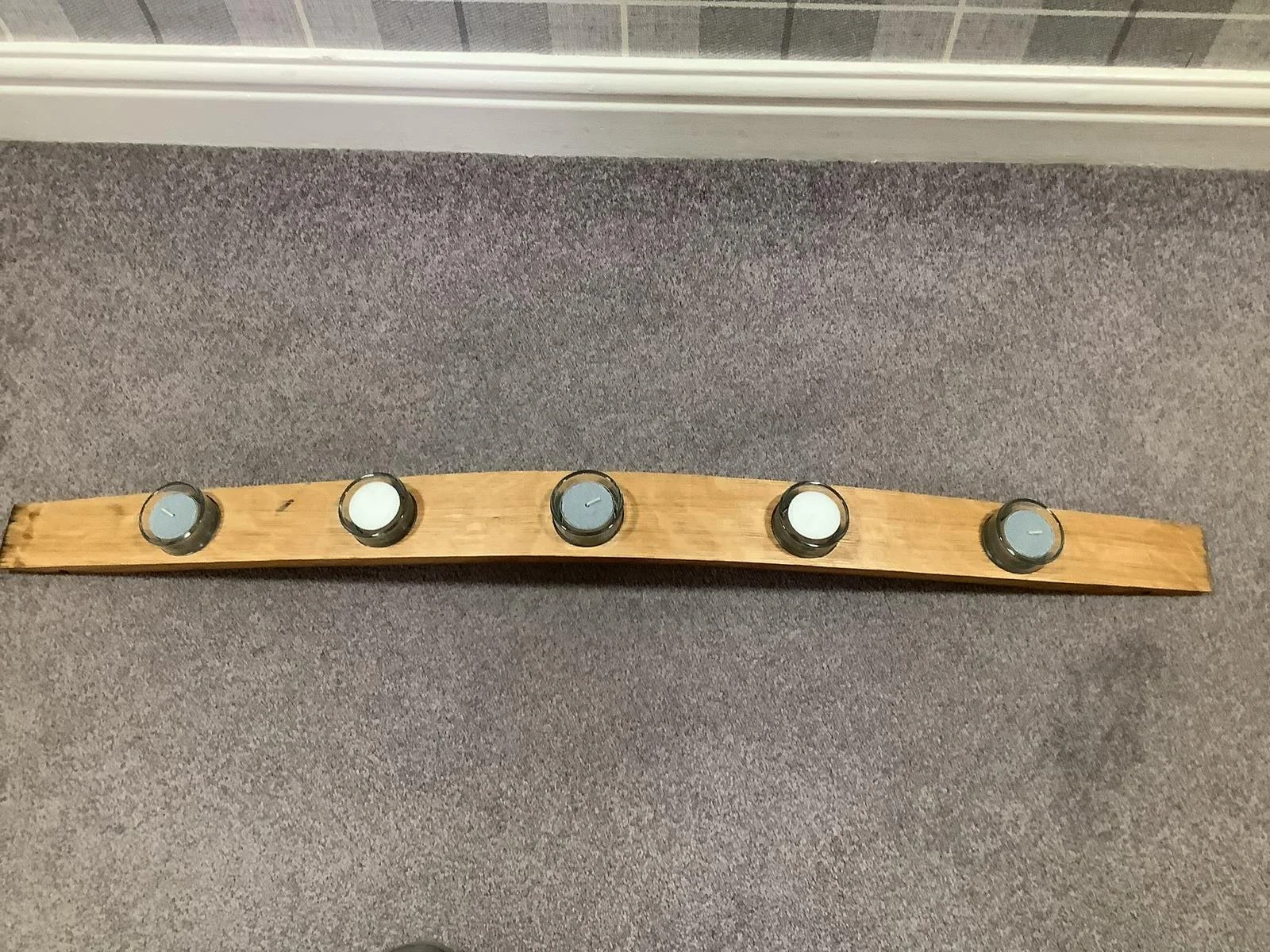Wooden plank with five tealight candles placed on a carpeted floor, with a wall and baseboard in the background.