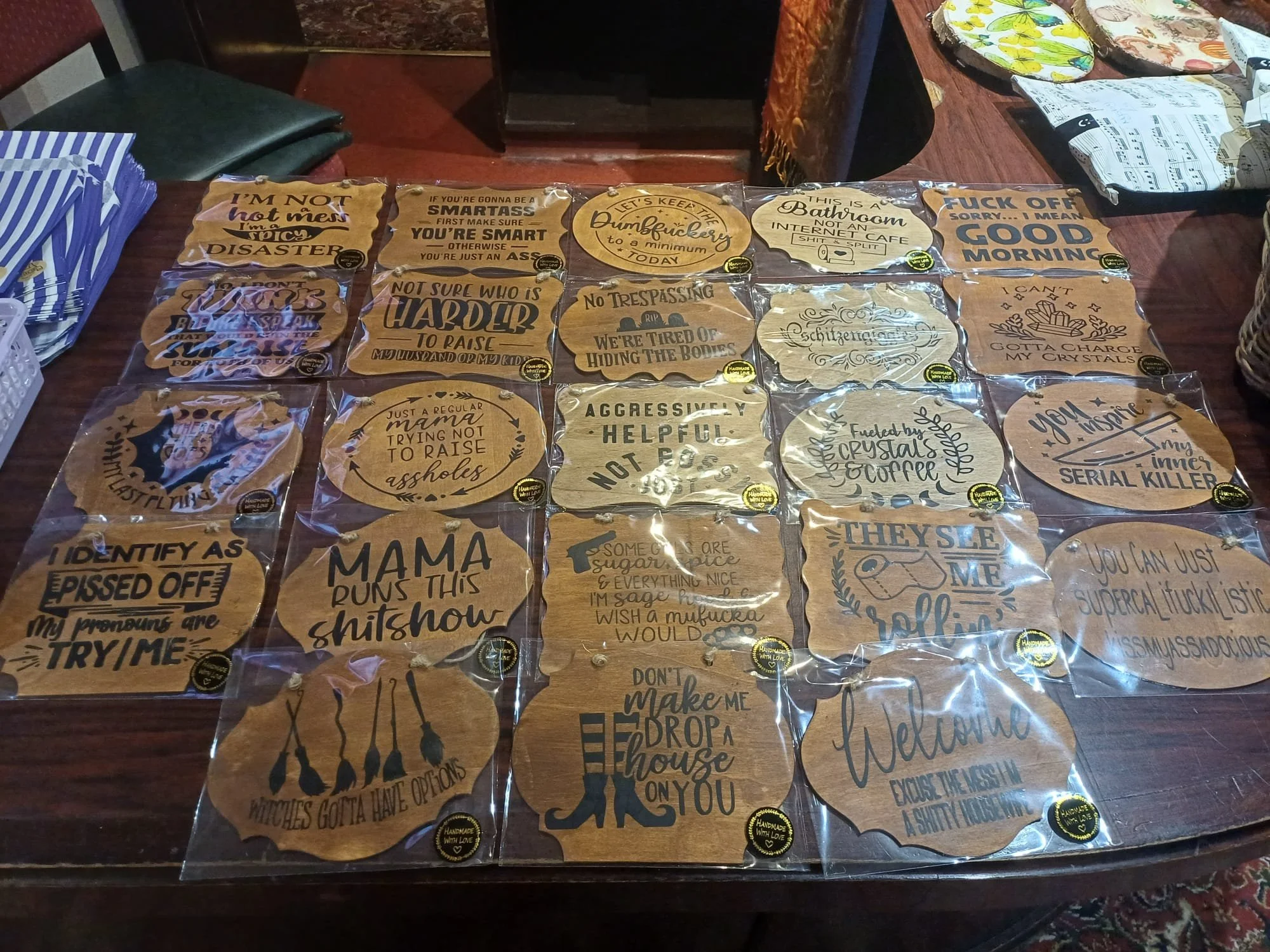 Collection of wooden plaques with humorous and sassy sayings on a table, some in plastic wrapping, with additional items and furniture around.