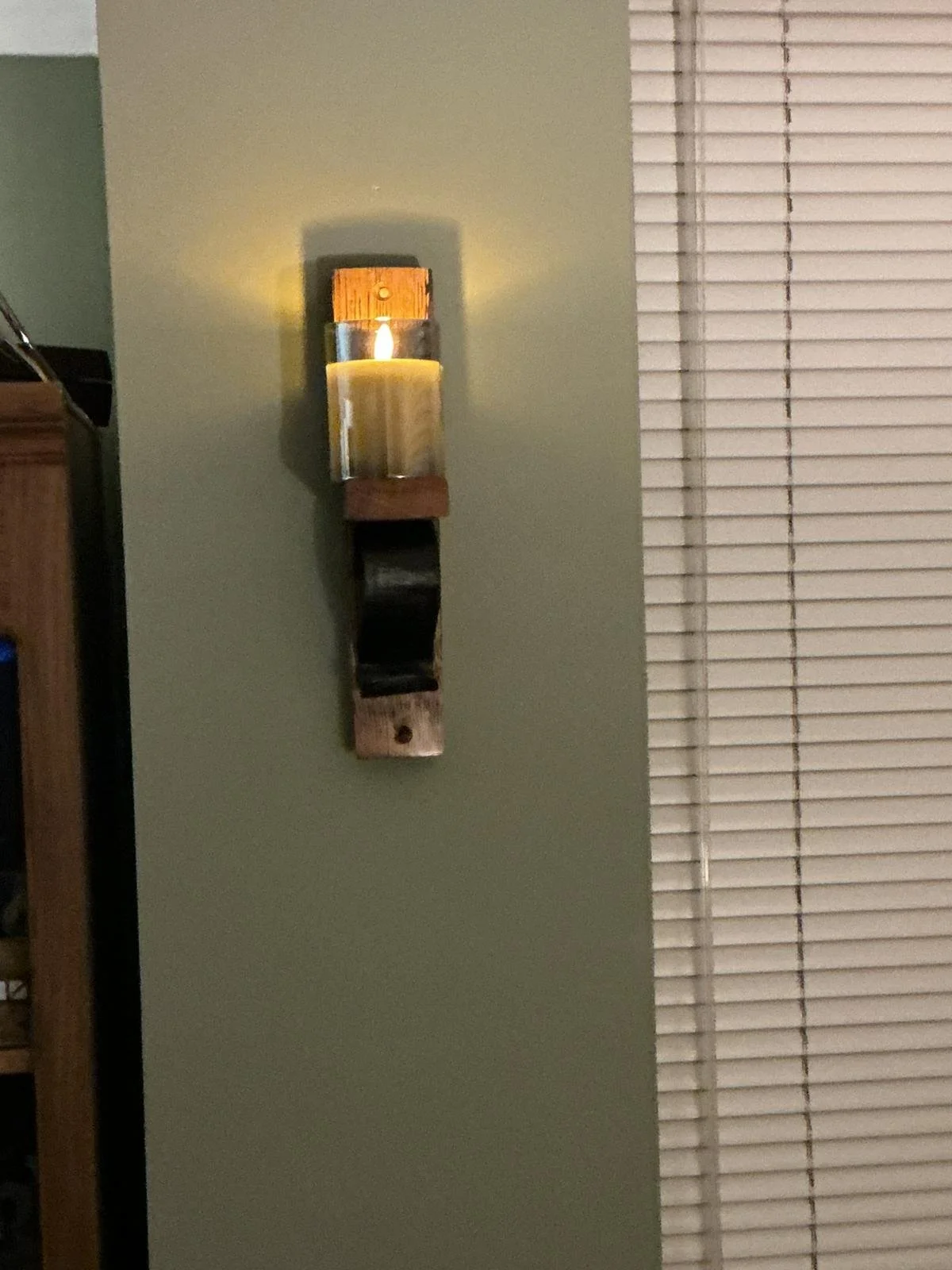 Wall-mounted candle holder with a lit candle casting a warm glow on the wall, adjacent to a window with closed blinds.