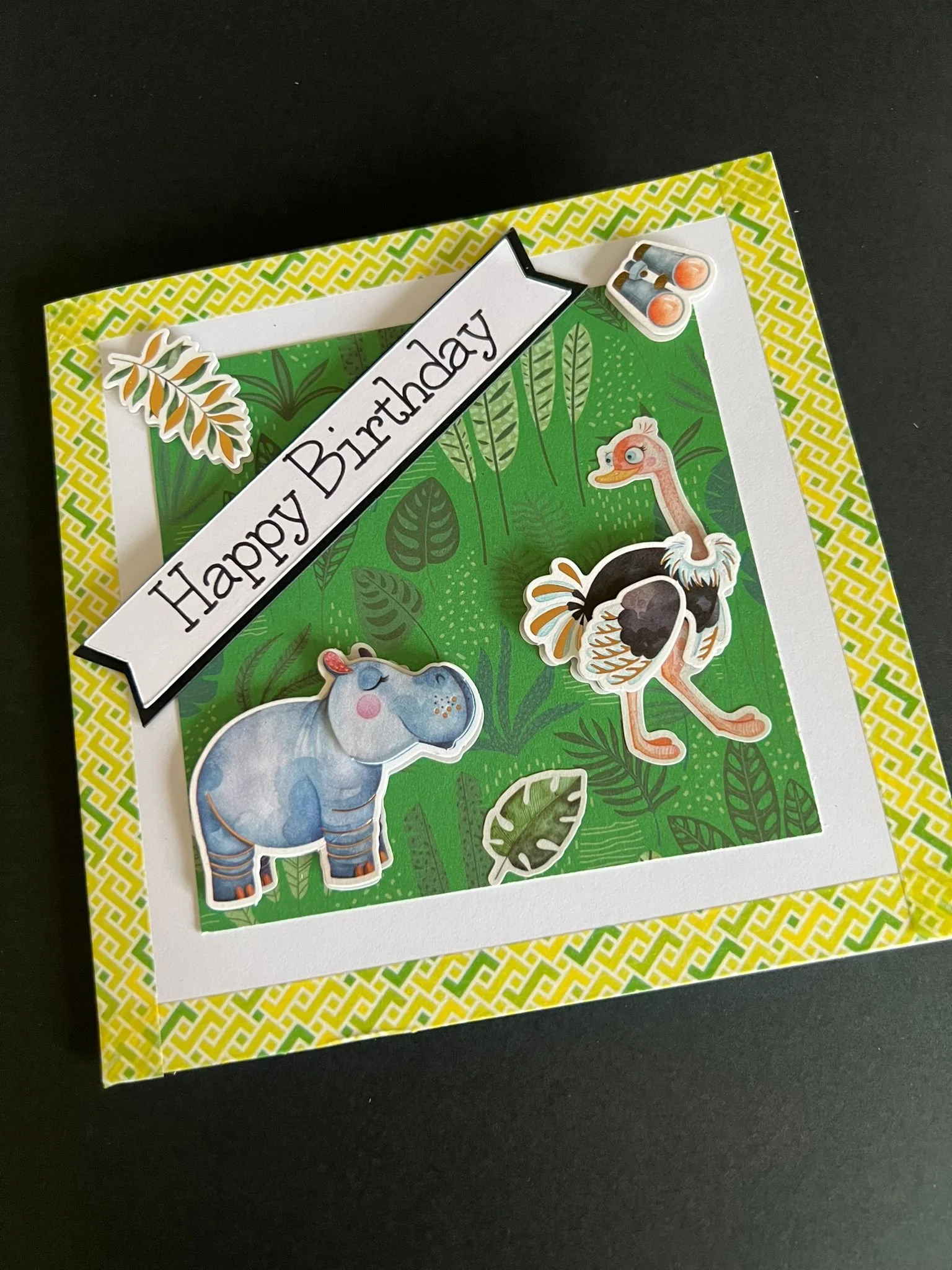 Colorful birthday card with jungle animals sticker set including a hippopotamus, a stork or flamingo, a giraffe, and foliage, with a banner reading 'Happy Birthday' at the top left.
