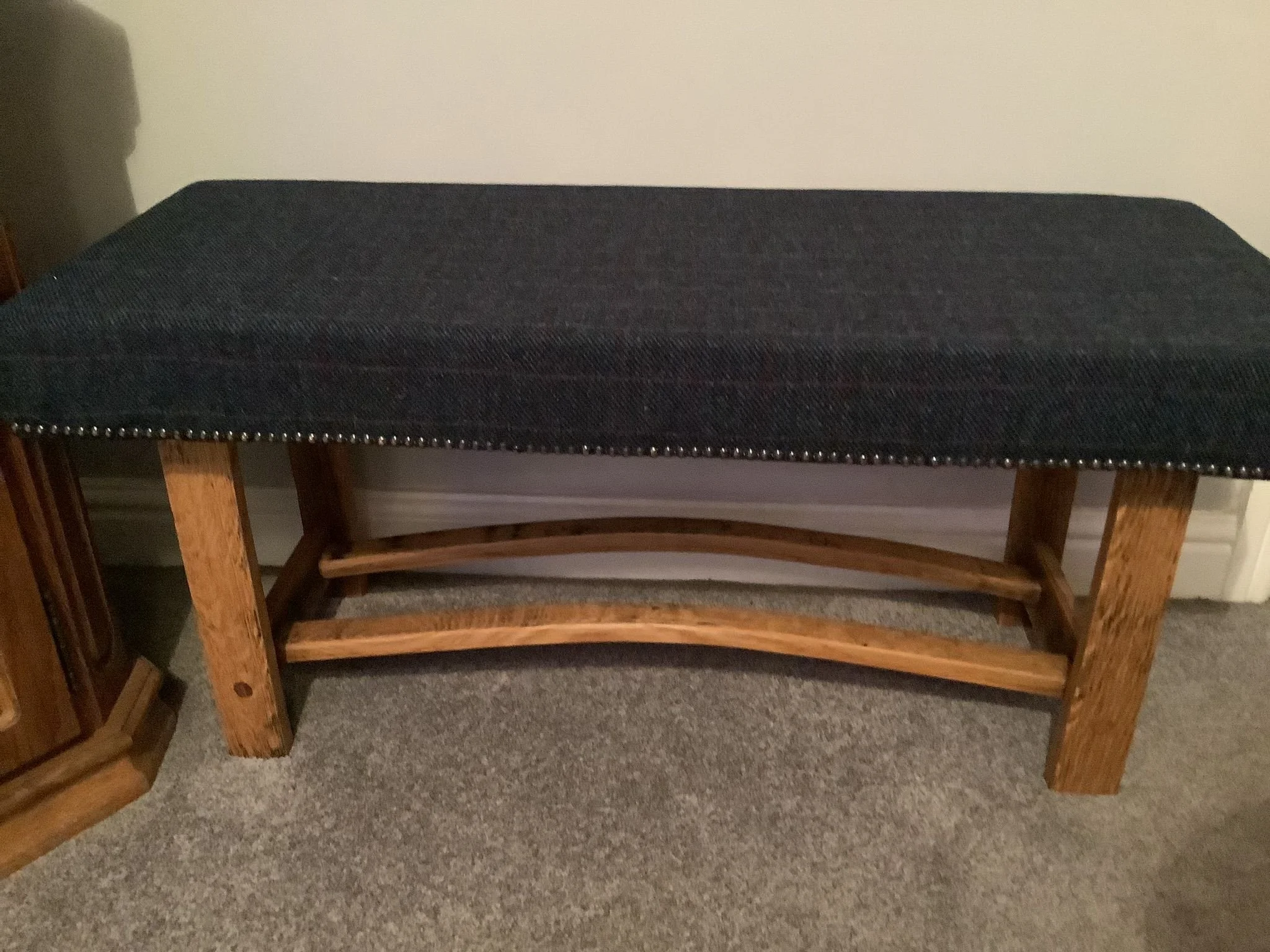 A wooden bench with a dark upholstered cushion on top, situated on a carpeted floor with a plain wall in the background.