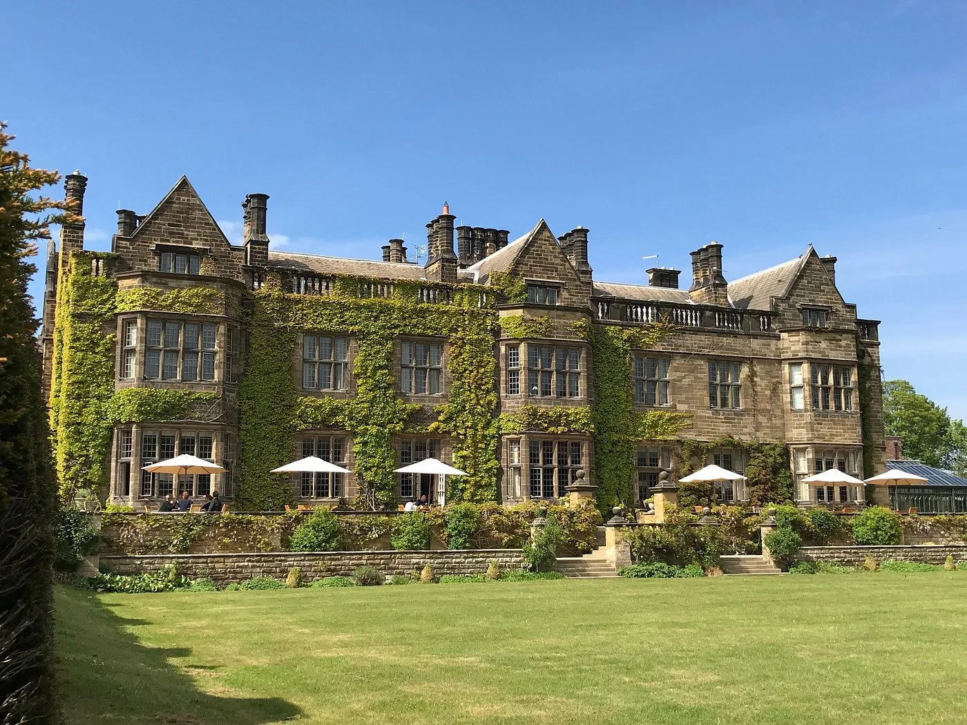 A large historic castle-like building with ivy growing on its walls, positioned behind a manicured lawn and stone steps. Several white patio umbrellas and people sitting at outdoor tables are visible on a sunny day, with a blue sky overhead.
