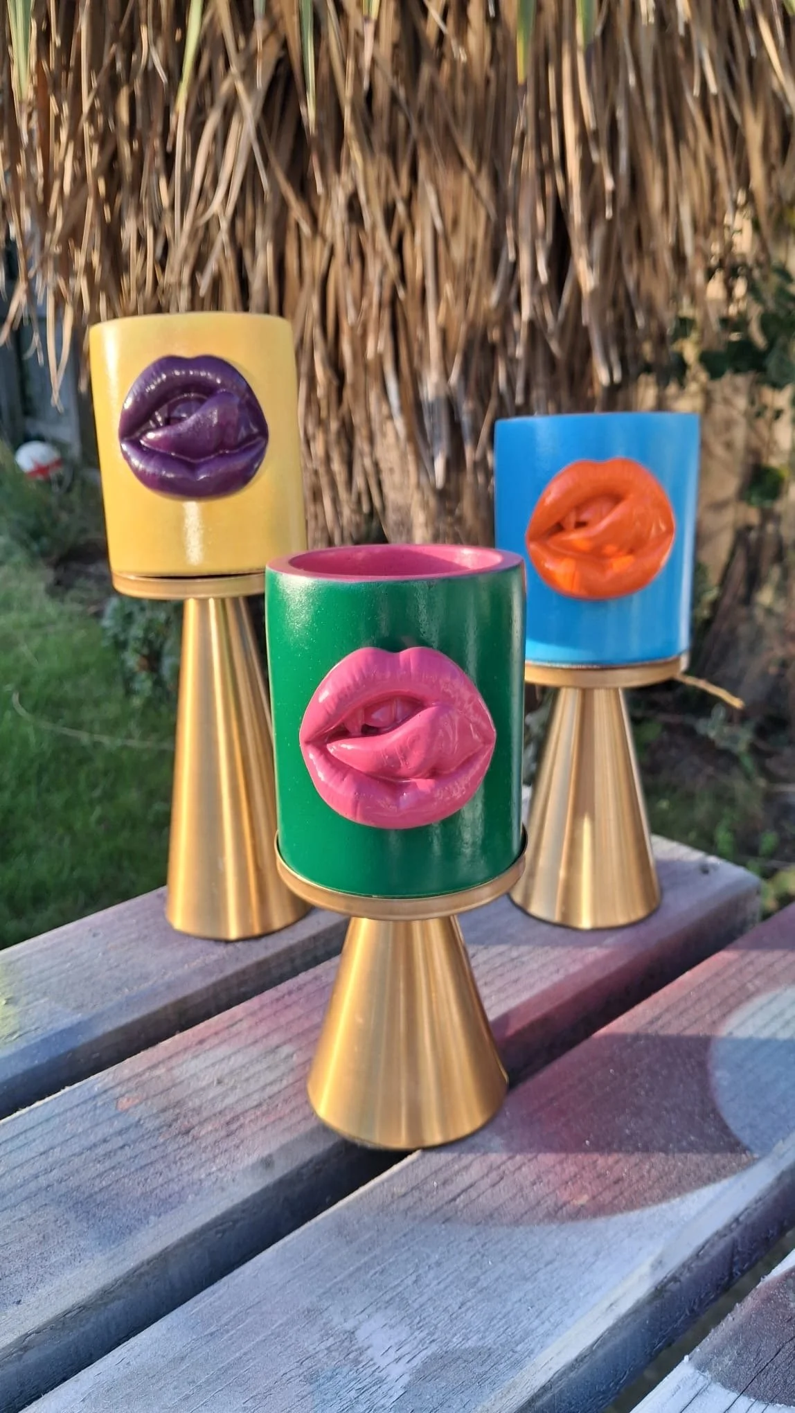 Colorful candles with lips-shaped designs on top, arranged on gold cone bases outdoors, near a wooden fence and grass.