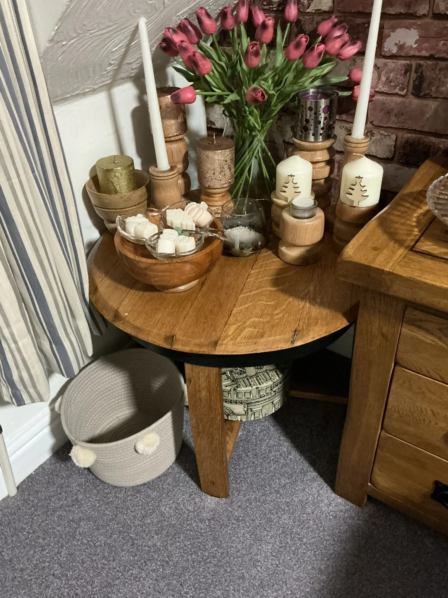 Side table decorated with pink tulips, candles, and holiday ornaments, with a basket underneath on carpeted floor.