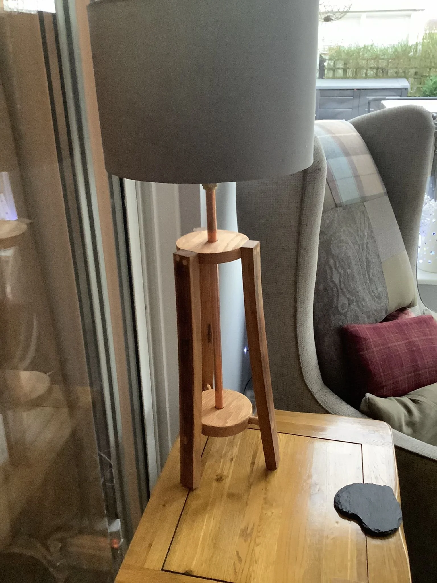 A wooden table lamp with a fabric lampshade placed on a wooden table next to a sofa with a plaid pattern and pillows.