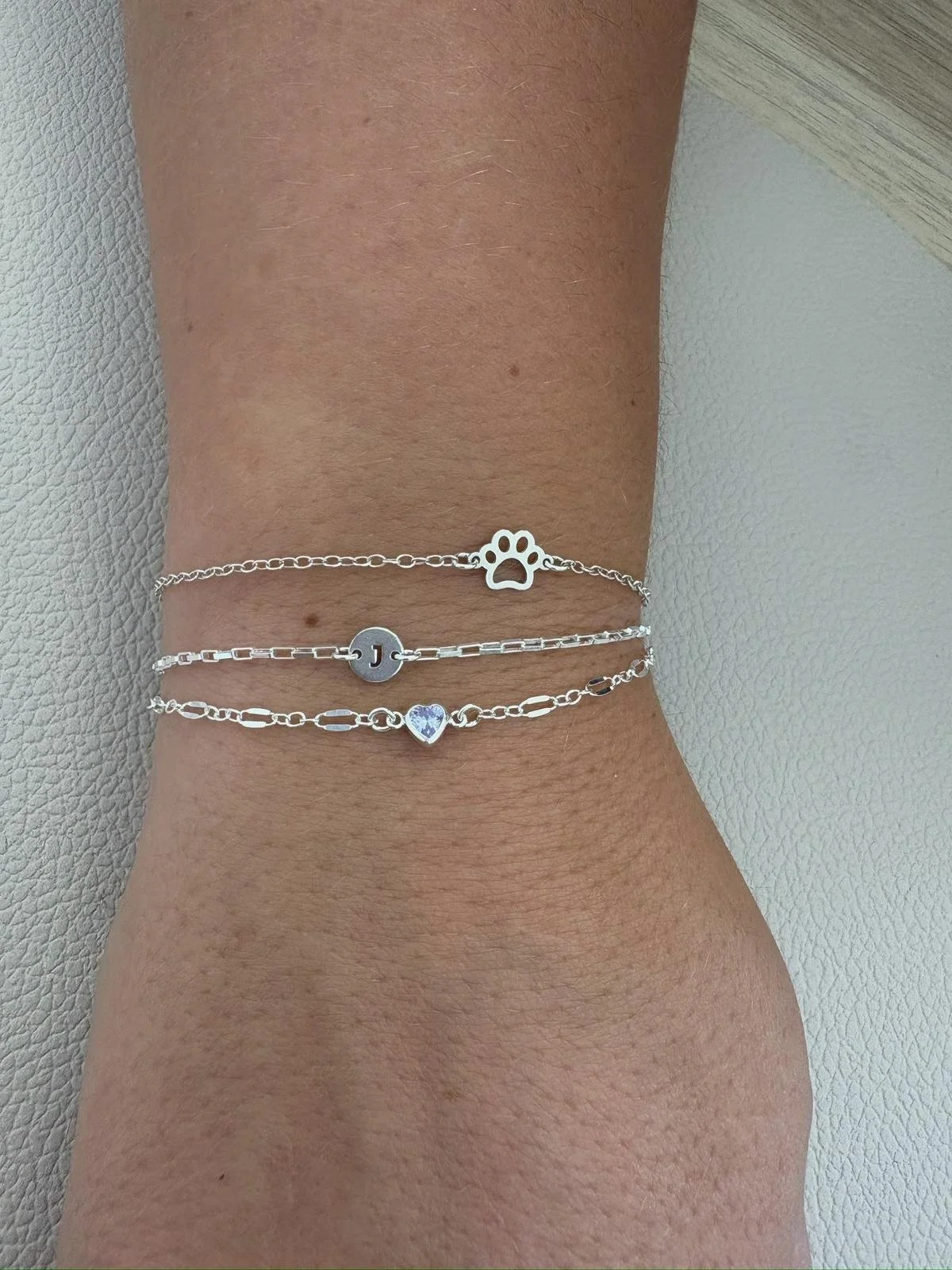 A close-up of a person's wrist wearing three silver bracelets with different charms: a paw print, the letter 'J', and a heart with an iridescent surface.