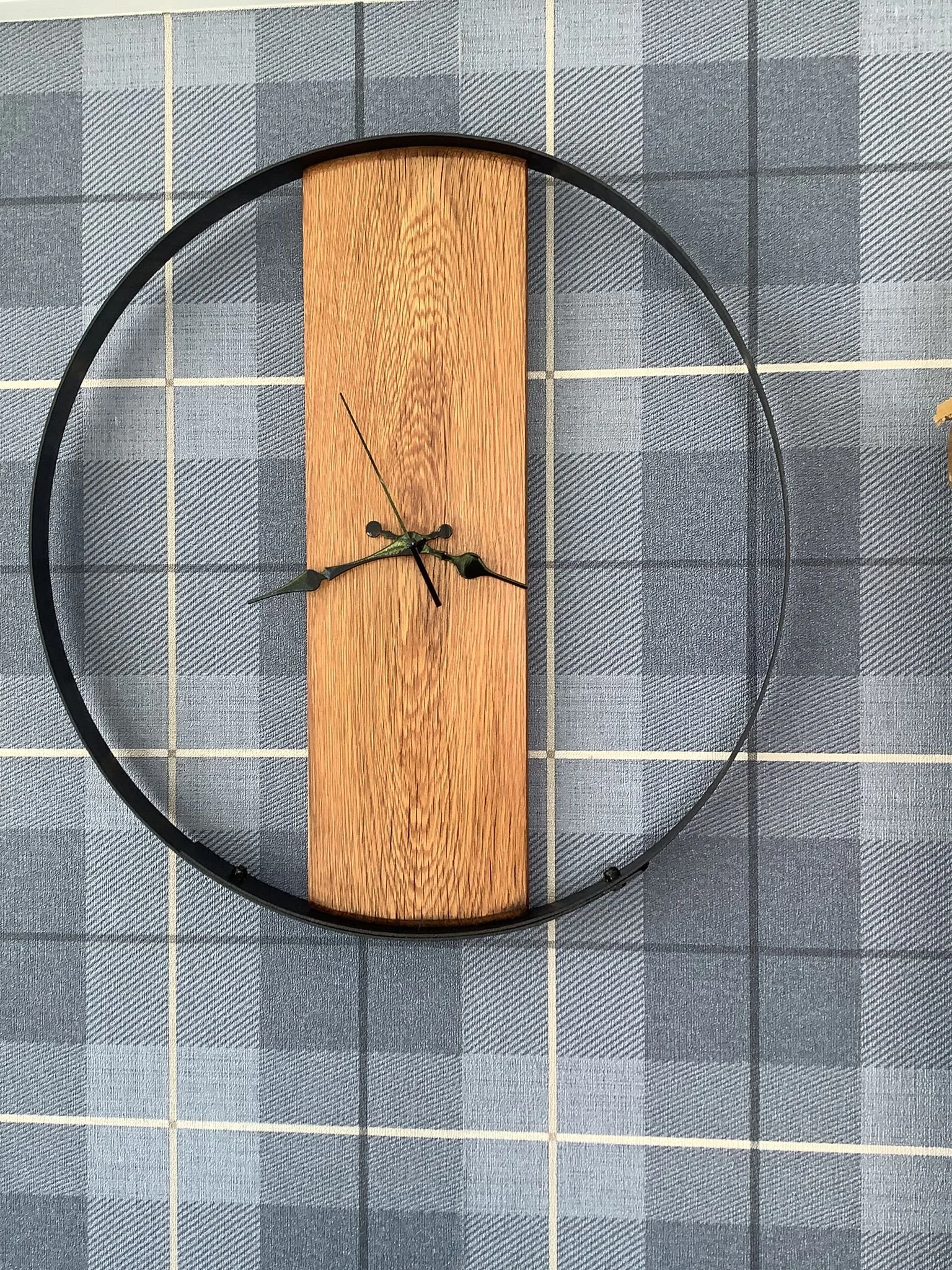 A modern clock with black rim, wooden center, and black hands, resting on a blue plaid fabric surface.