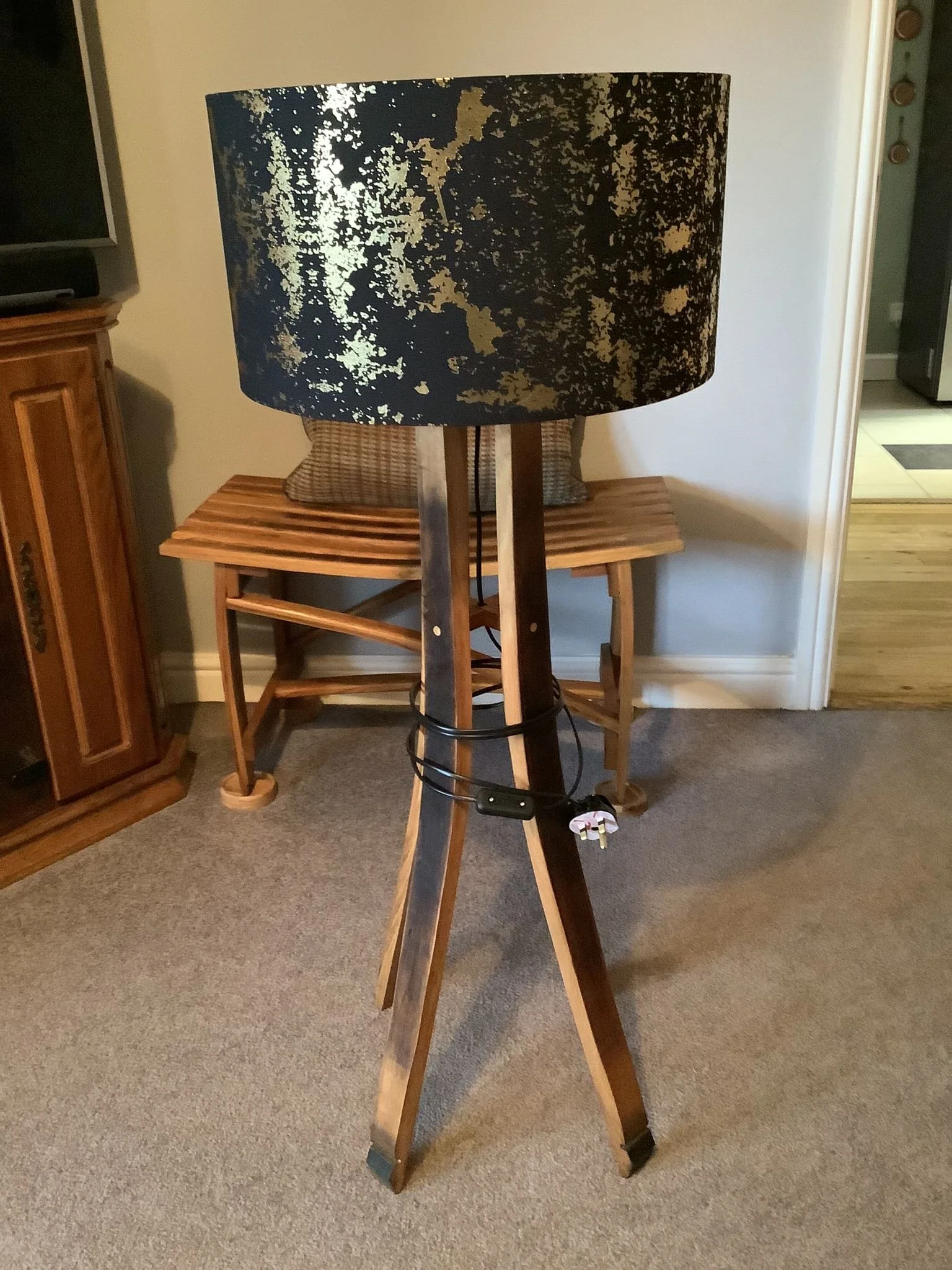 A floor lamp with a black and gold patterned lampshade, wooden tripod base, and electrical cord with plug, positioned on carpeted floor near a wooden bench and wall.