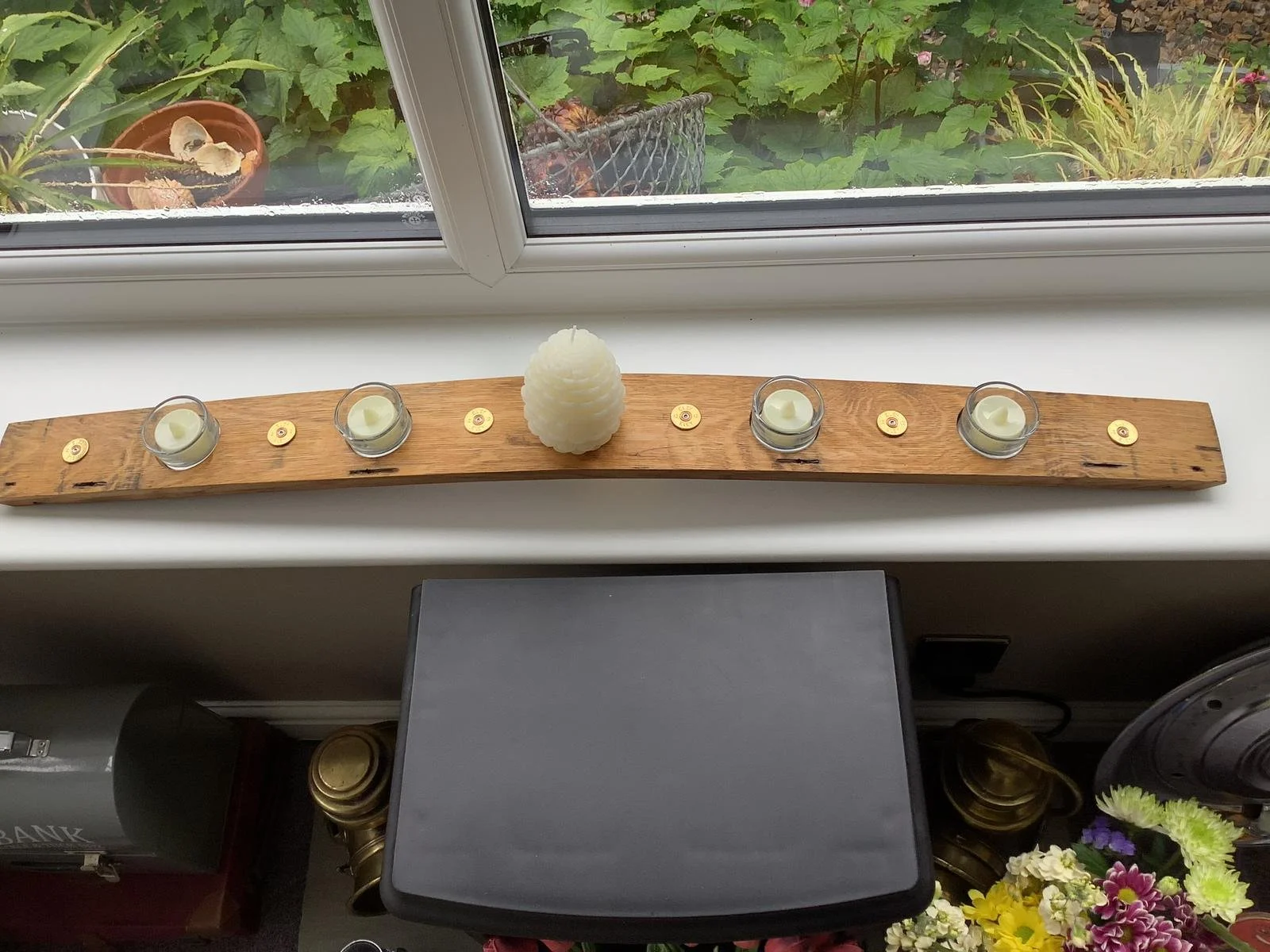 A wooden candle holder with five white tealight candles and one large white decorative candle on a windowsill, overlooking a garden with green plants and pink flowers outside.