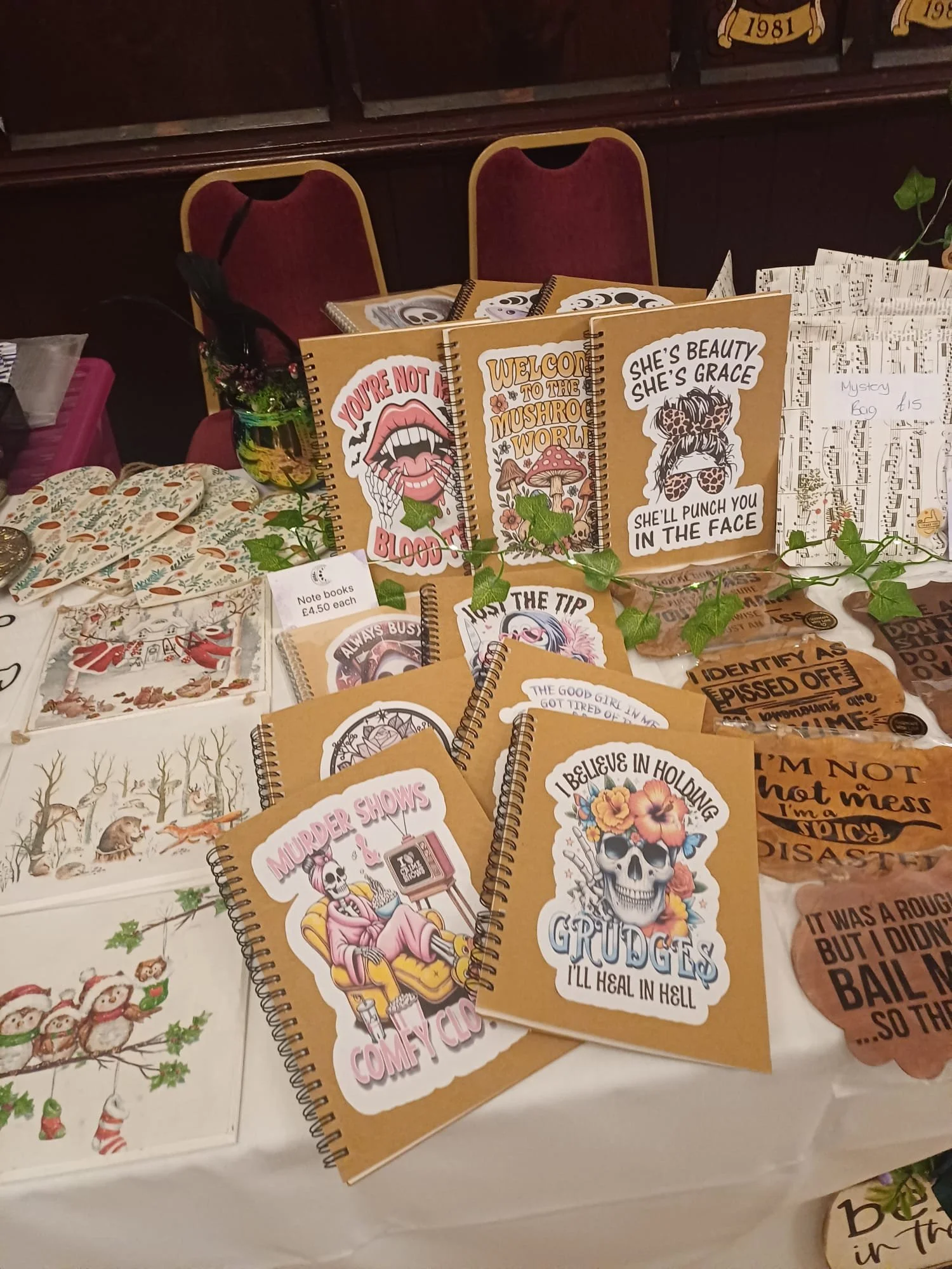Table display of humorous notebooks with tattoo-inspired designs and quotes, including phrases like 'You're not that blood,' 'Welcome to the mushroom world,' 'She's beauty, she's grace, she'll punch you in the face,' and 'I believe in holding grudges till heal in hell.'