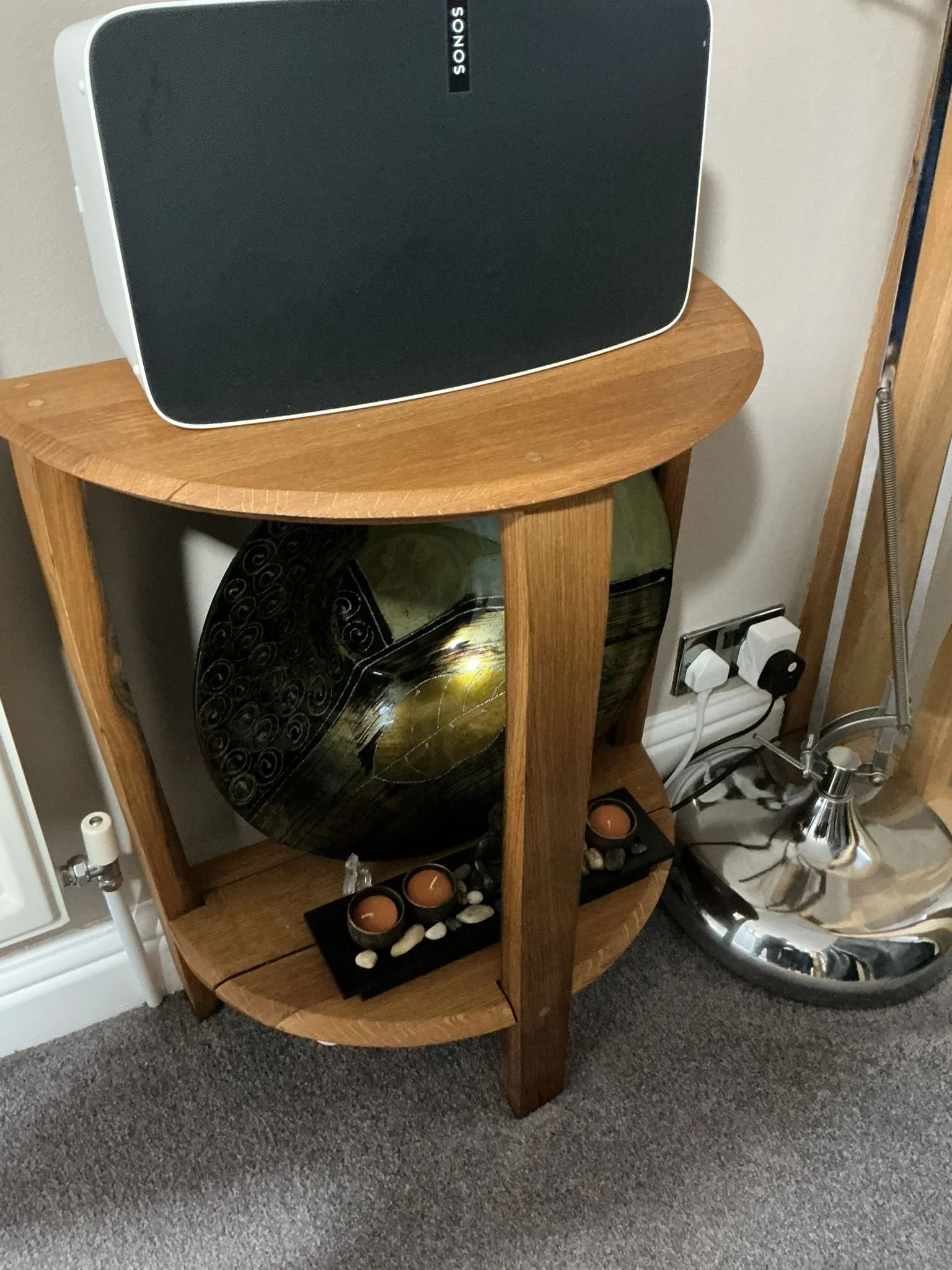 A wooden corner shelf with a black and gold decorative plate, three candles, and small stones. A Sonos speaker is on top of the shelf, and a chrome lamp is nearby.