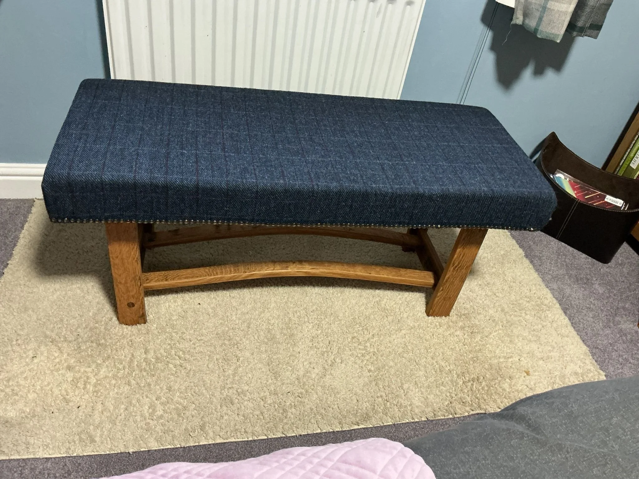 A wooden bench with a dark blue cushioned top placed on a beige carpet. There is a greenish-gray wall behind the bench, a radiator, and a pocket organizer with magazines on the right side.