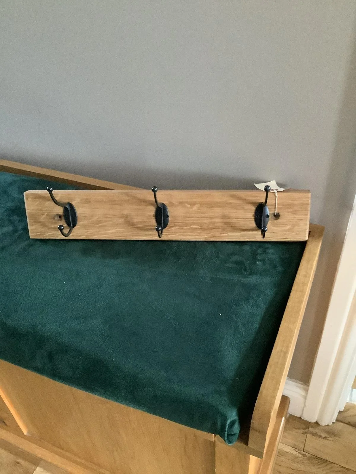 Wooden rack with four black hooks attached, mounted on the side of a piece of furniture with green fabric covering.