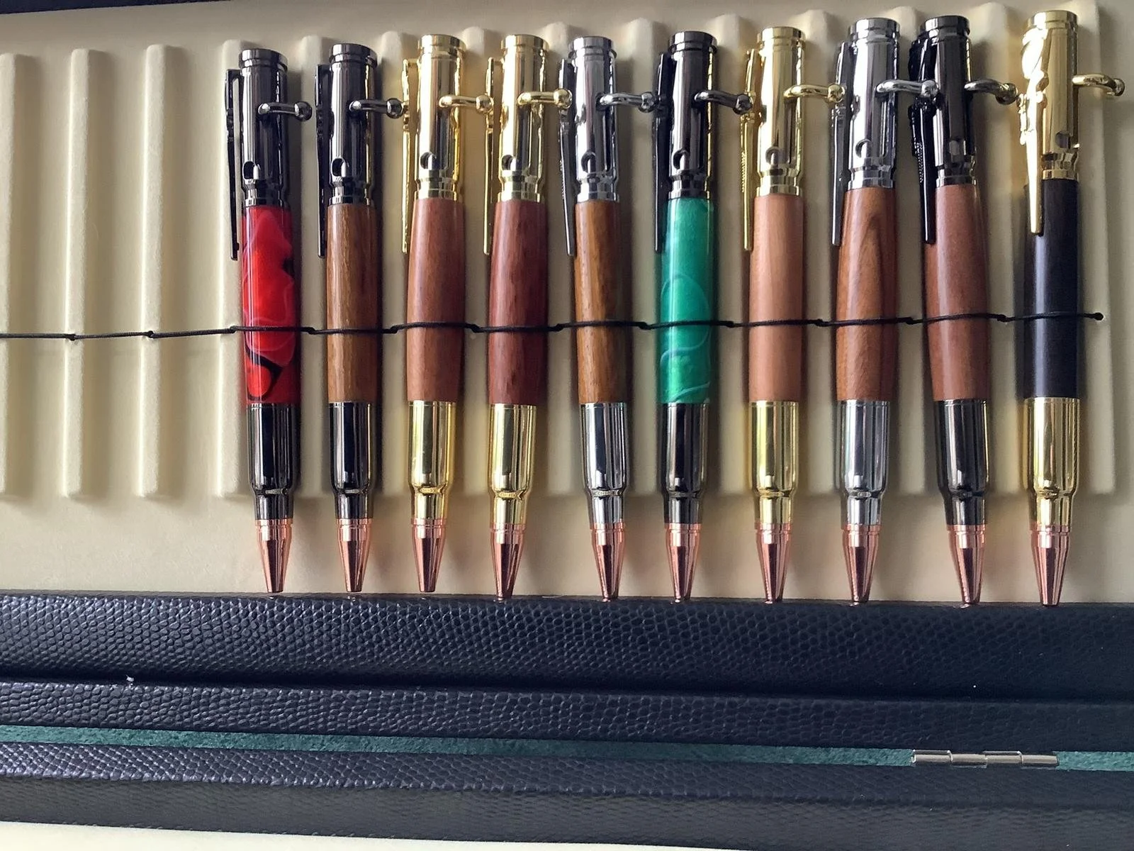 A collection of ten ballpoint pens with wooden barrels and gold, black, or silver accents, arranged in a row on a foam-lined case.