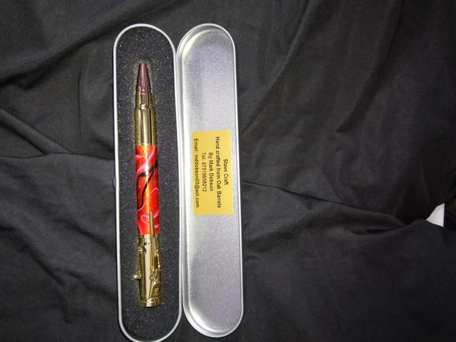 A decorative pen with a red and black swirling design in a silver tin case with a yellow warning label.