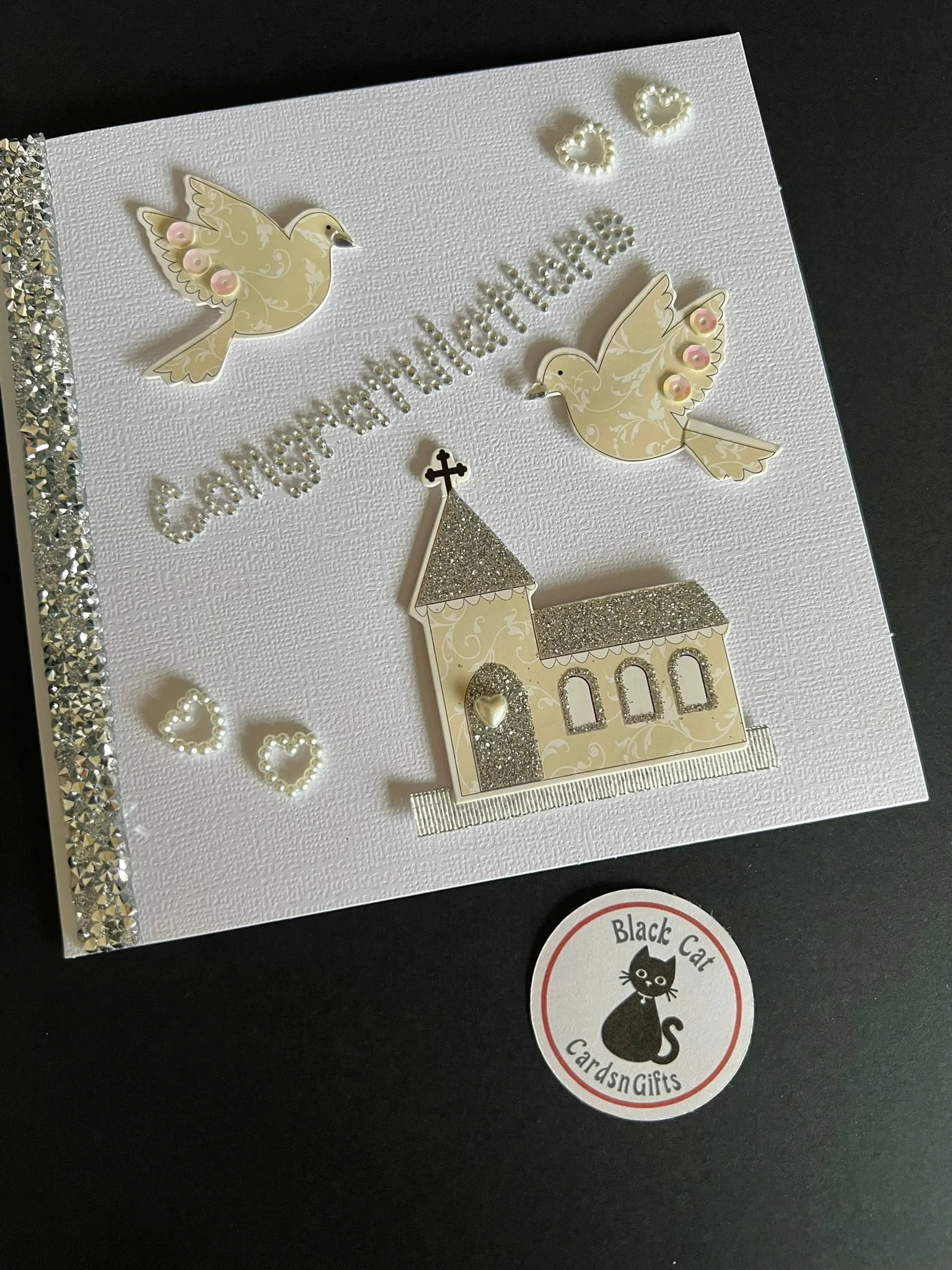 White greeting card with two decorated doves, a church, and pearl accents, featuring the word 'Congratulations' in glitter, with a logo sticker at the bottom right reading 'Black Cat Cards and Gifts'.