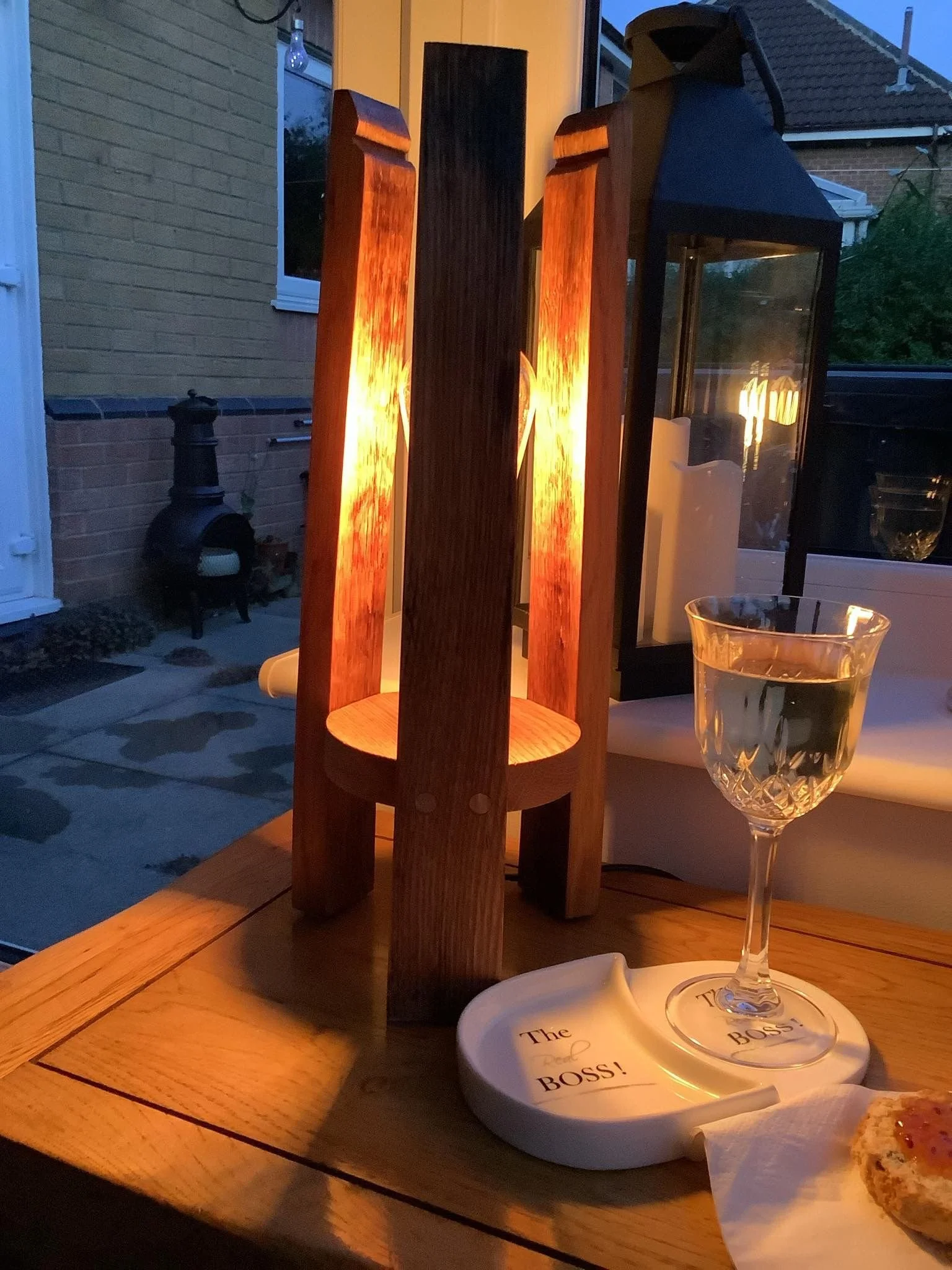 A lit candle lantern on a wooden table, a glass of white wine, a paper towel, and a coaster with the text 'The Boss!' on it, with a piece of bread or pastry nearby. The background shows a patio and house exterior at dusk.