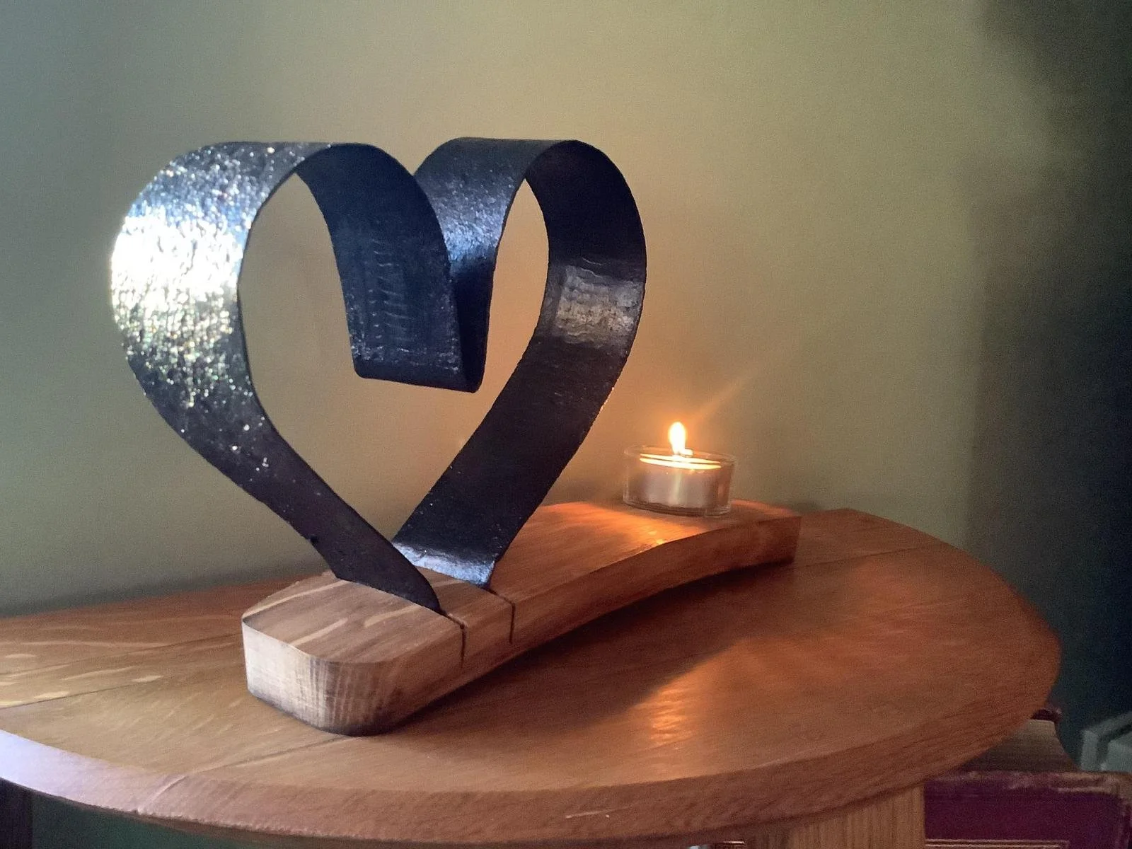 A decorative metal heart sculpture on a wooden base, with a small lit tealight candle beside it on a wooden surface.