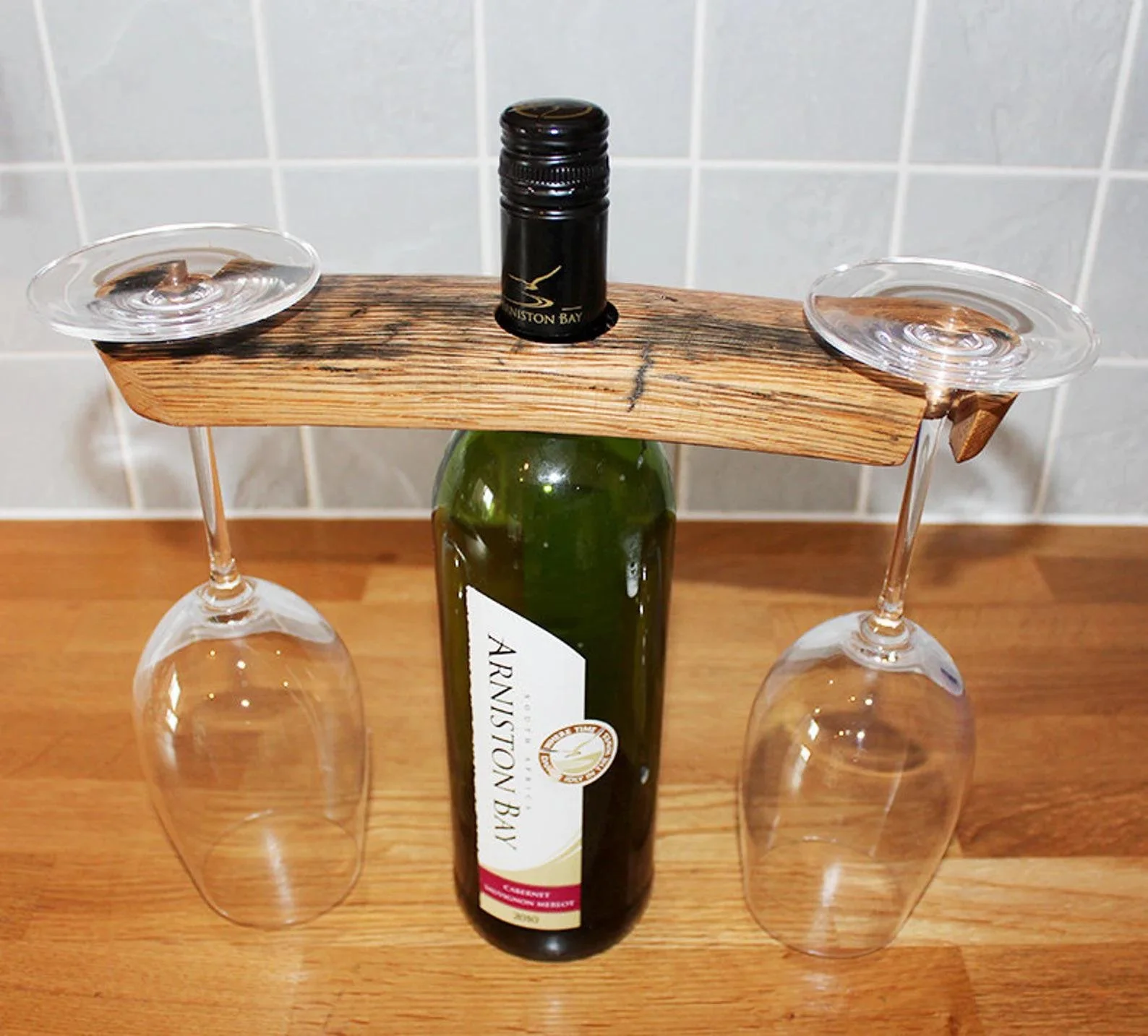 A wine bottle balanced on a wooden plank held between two upside-down wine glasses on a wooden surface.