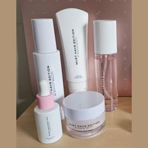 A collection of five skincare or cosmetic products, including bottles and jars, with labels reading 'MUST HAVE EDITION' and 'FACESOURCE' on a wooden surface against a pink background.