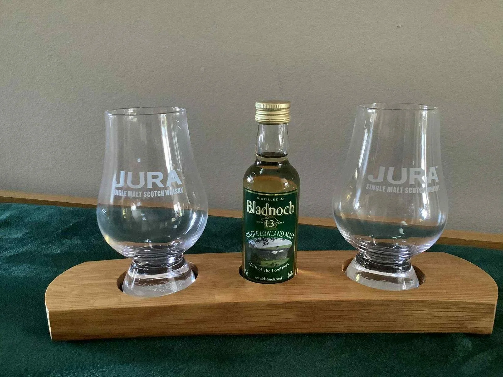 Two empty whisky glasses with JURA Single Malt Scotch Whisky logo on a wooden serving board, with a small bottle of Bladnoch Single Lowland Malt Whisky between them.