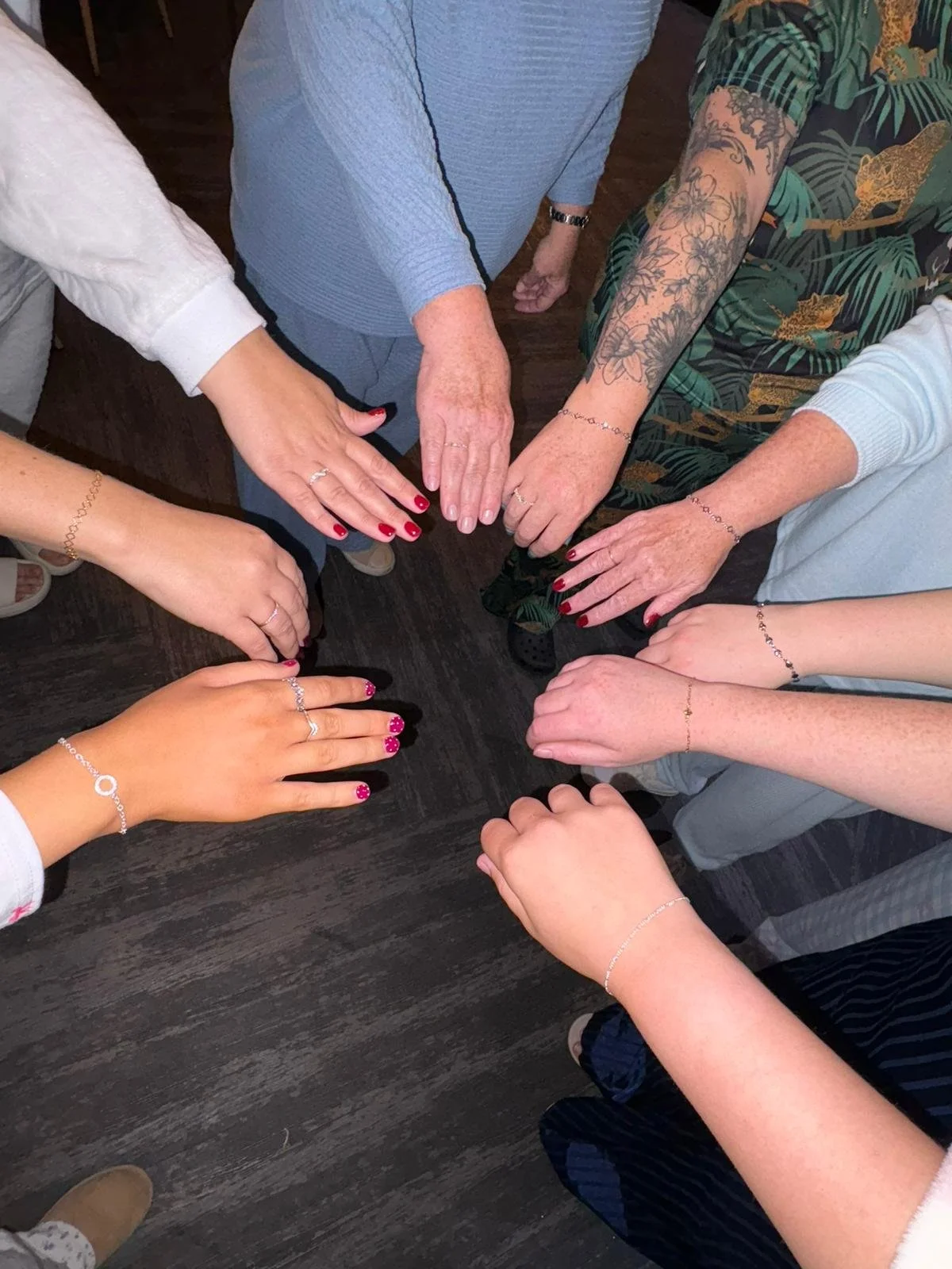 Multiple women with various skin tones and tattooed arms gather around a table, touching hands and displaying jewelry, including rings, bracelets, and nail polish in pink and red.