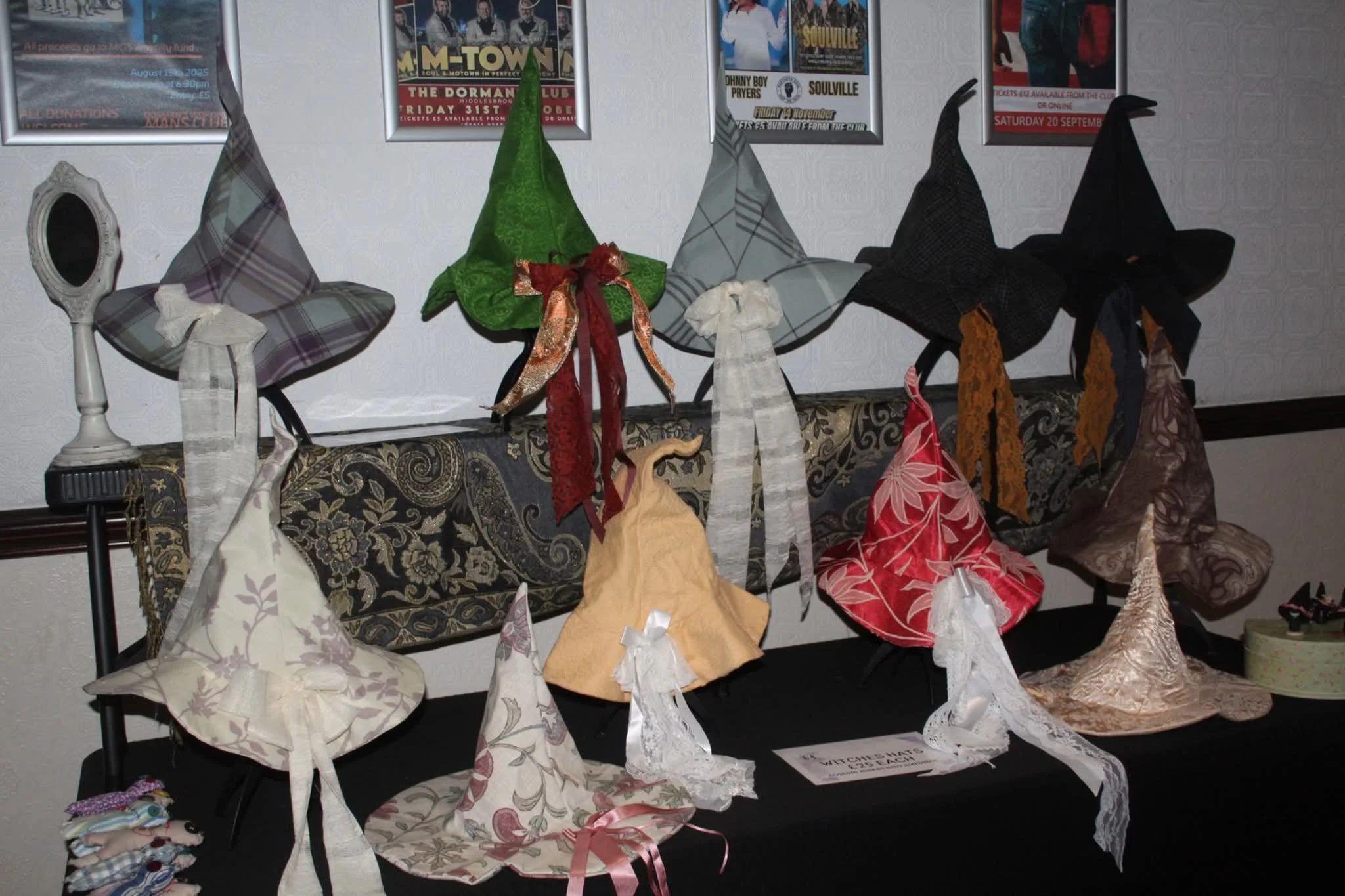 A display of handmade witch hats in various colors and fabrics arranged on a black table with some hats in the background. Posters on the wall may be seen behind the display.