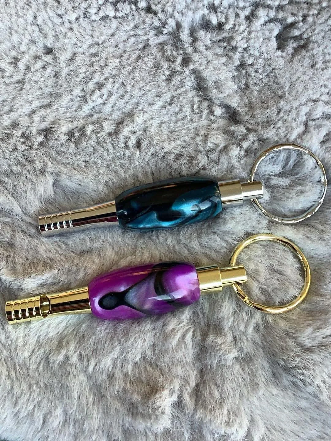 Two metal keychains, one with a purple and black marbled handle and one with a blue and black marbled handle, resting on a soft, furry surface.