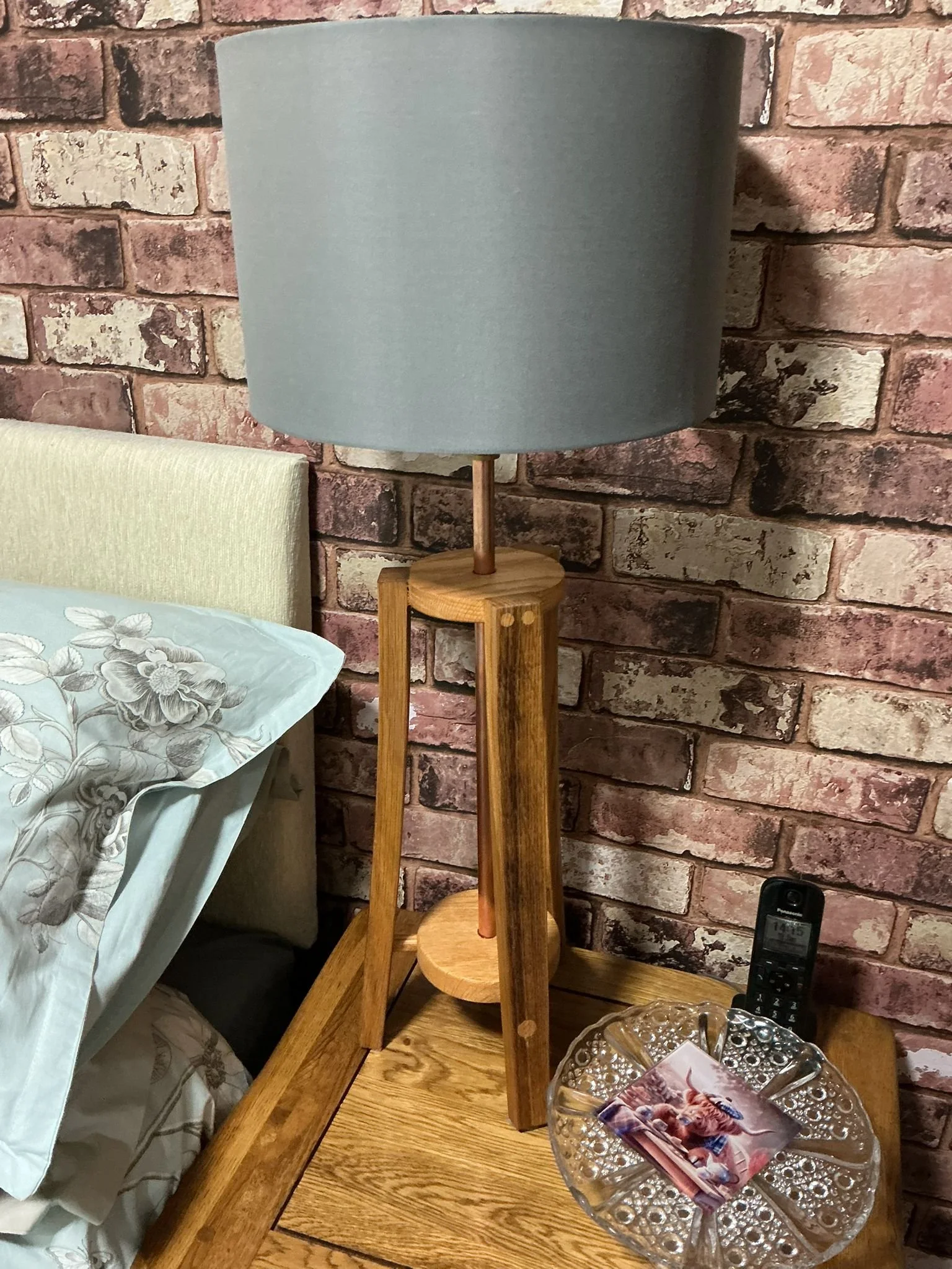 A wooden floor lamp with a large gray fabric lampshade on a wooden side table next to a bed with a floral pillow, against a brick wall. There is a glass dish with a photograph and a cordless phone on the table.