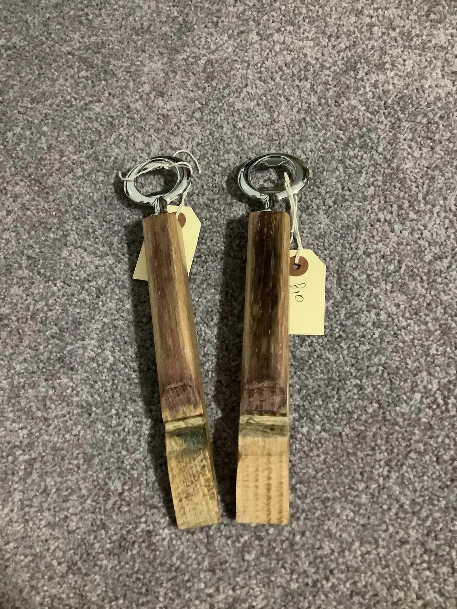 Two wooden-handled fire pit pokers with metal loops at the top, lying on a gray carpet. Each has a yellow price tag attached.
