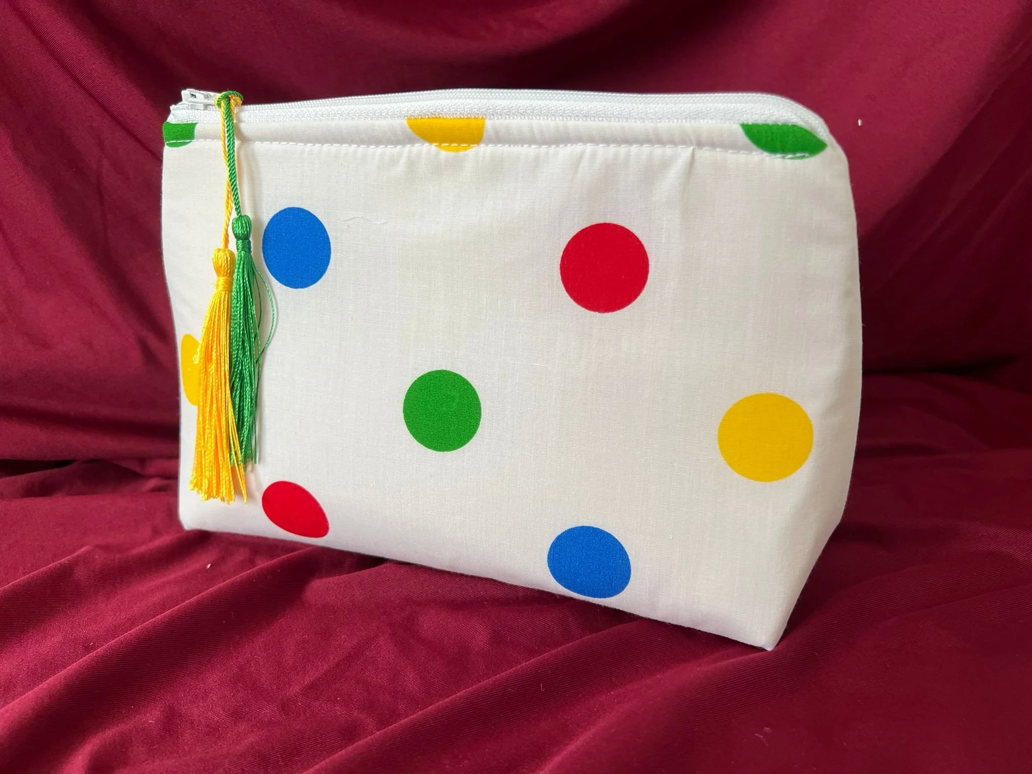 A rectangular fabric zippered pouch with a white background decorated with colorful polka dots in red, yellow, green, and blue, placed on a red velvet surface.