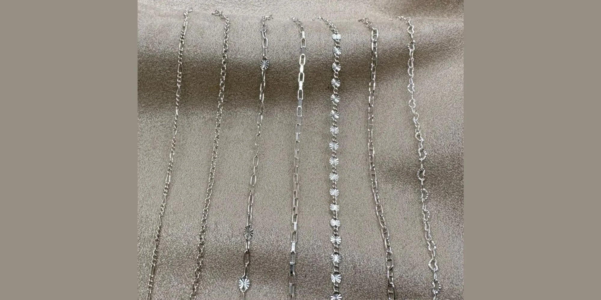 Six silver necklaces with various chain designs are laid out on a beige fabric surface.