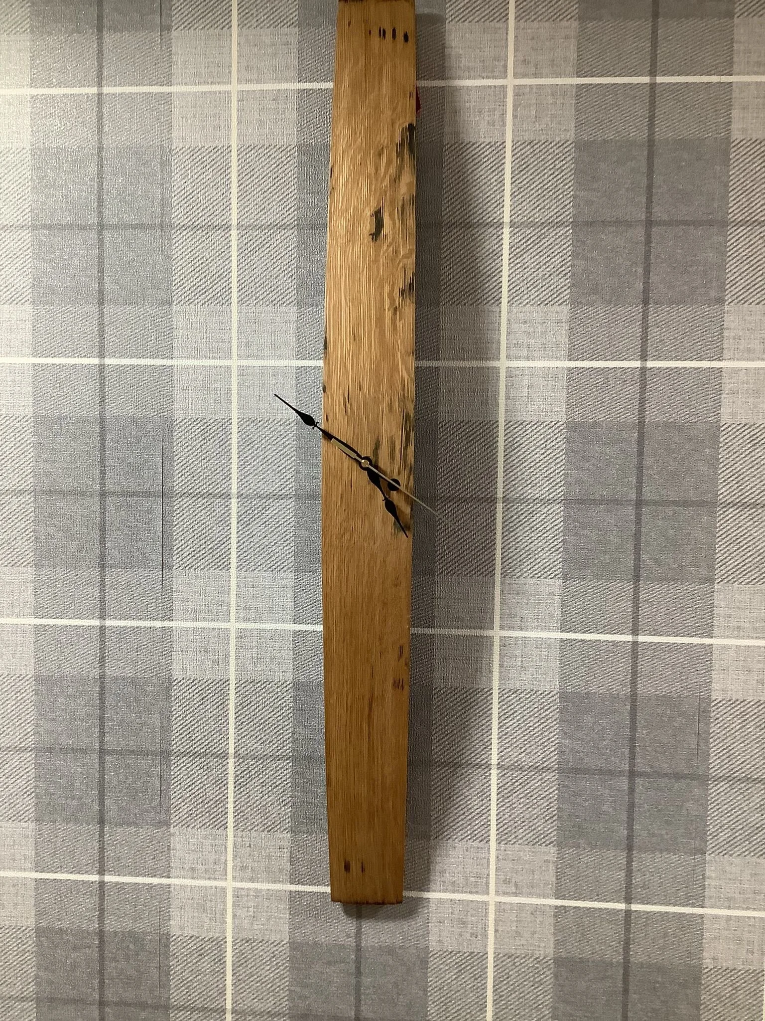 Clock with a minimalist design mounted on a long, vertical wooden plank against a checkered fabric background.