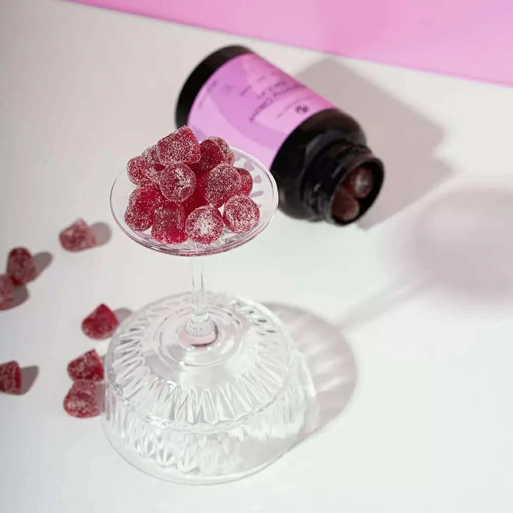 A glass dish with sugar-coated red gummy candies on a clear pedestal, a black bottle with a pink label lying on its side, spilling some candies, on a white surface with pink background.