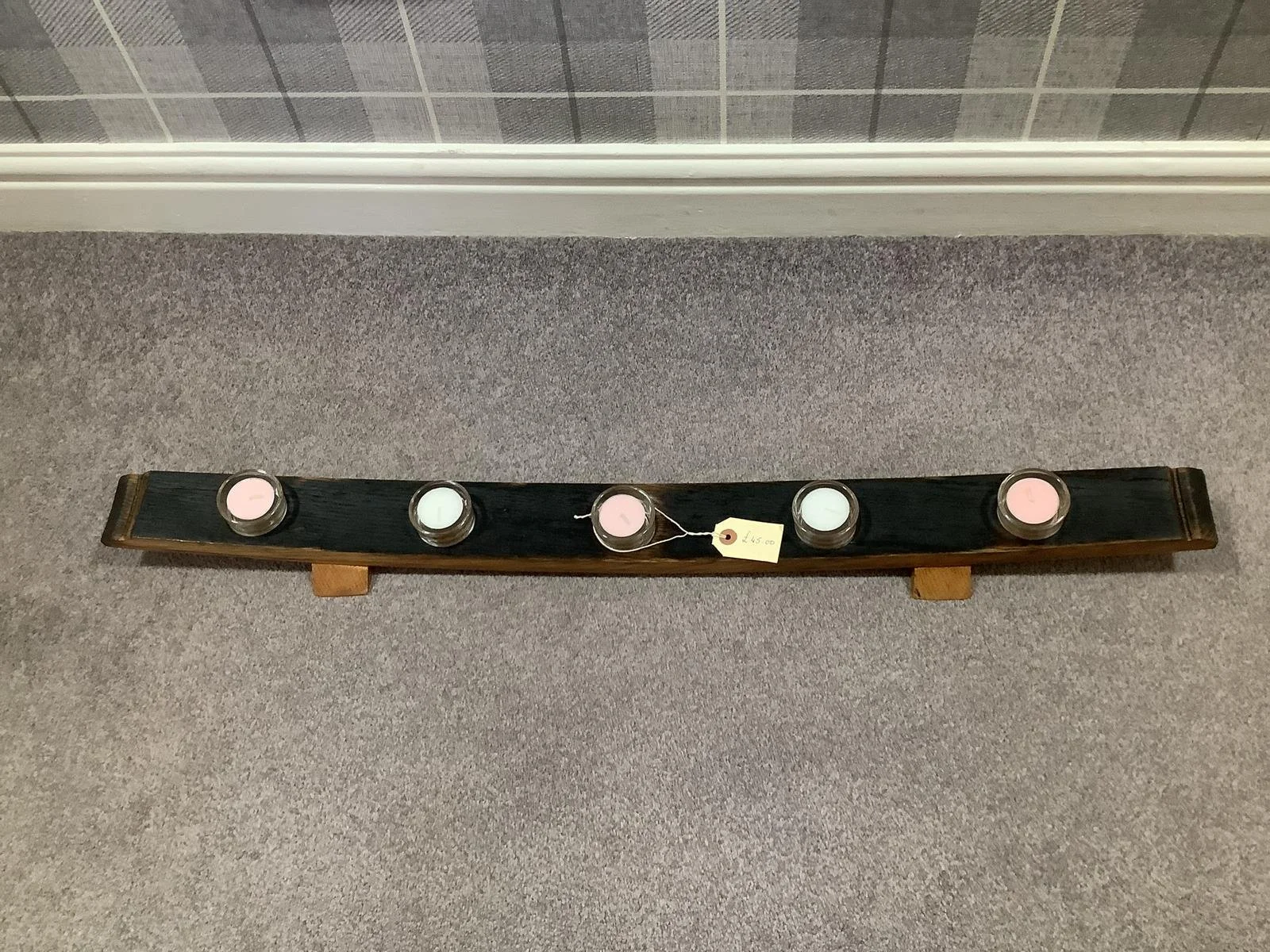 A wooden stand holding five candles, with three pink and two white, placed on a beige carpeted floor near a wall with wallpaper.