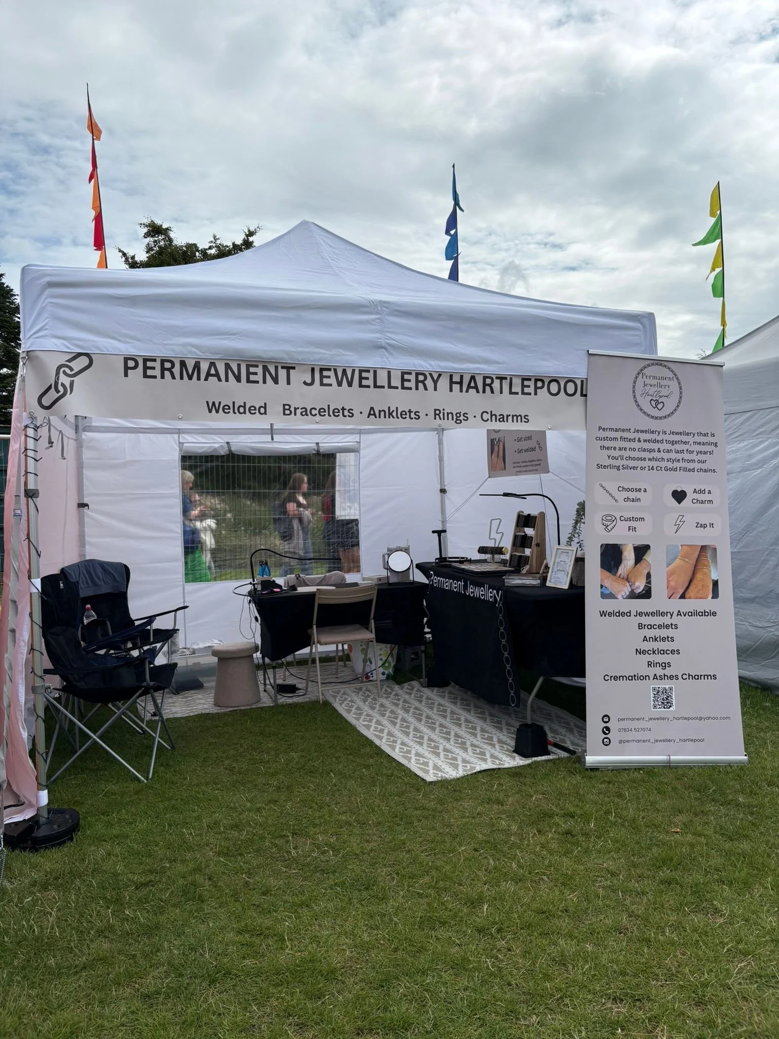 A white event tent with a sign reading "Permanent Jewellery Hartlepool" offering welded bracelets, anklets, rings, and charms. The tent has a table with jewelry tools and display items, a black chair, and a patterned rug on the grass outdoors. Flags 