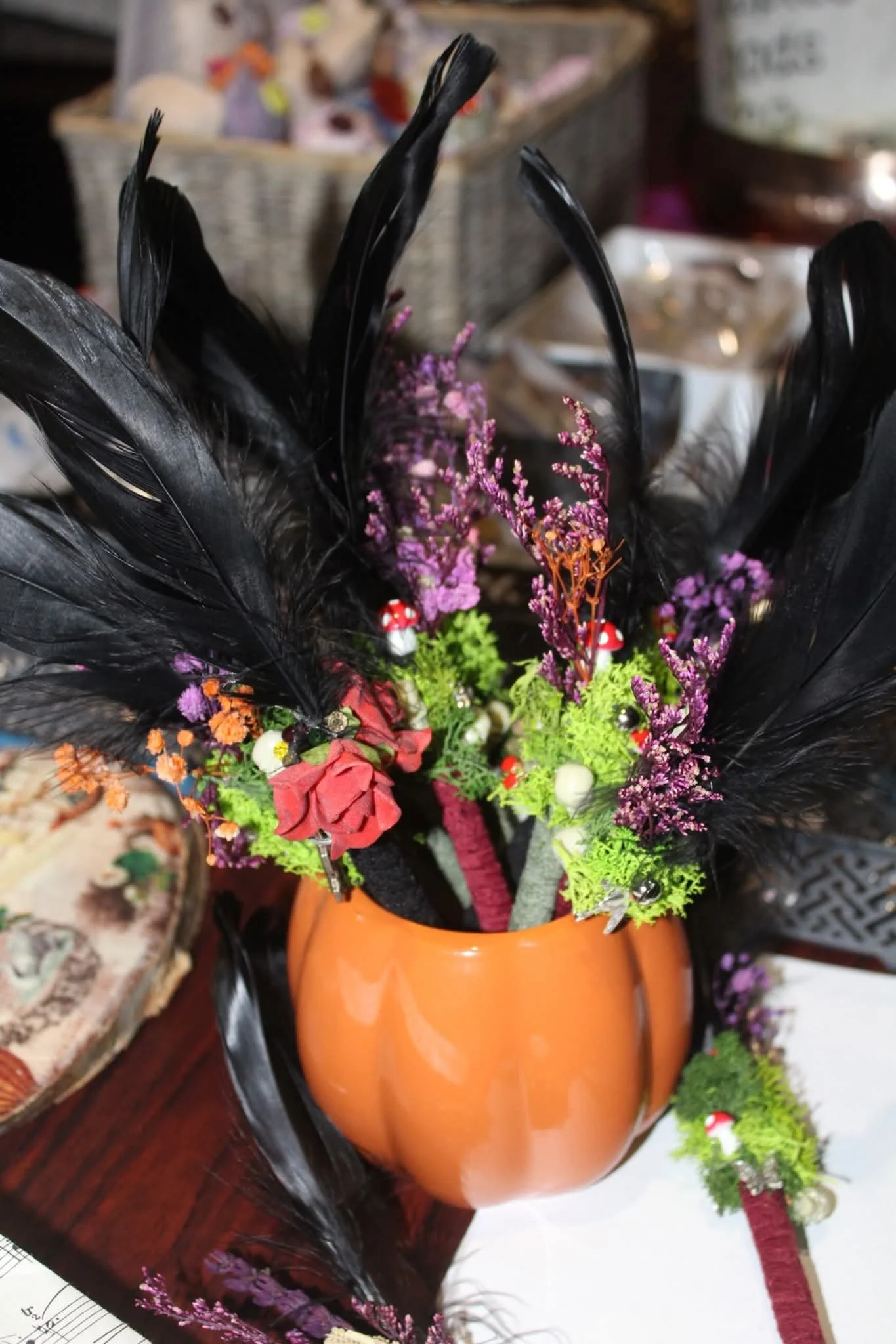 A decorative arrangement of artificial flowers, including black feathers, pink and purple flowers, mini mushroom ornaments, and green foliage in an orange ceramic mug.