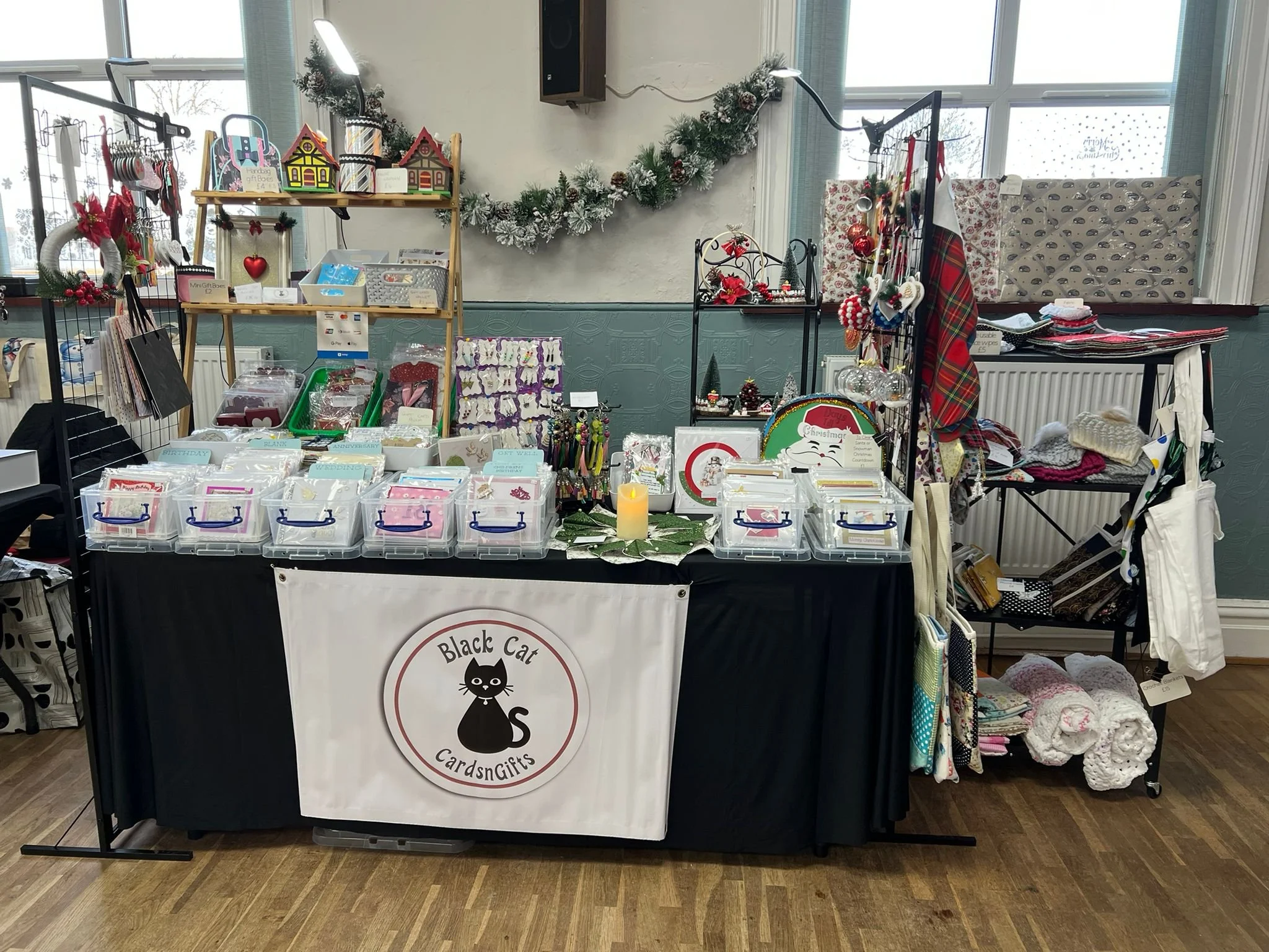 Christmas-themed craft and gift shop display with various holiday cards, ornaments, and decorations at a booth labeled 'Black Cat Cards & Gifts' in an indoor setting.