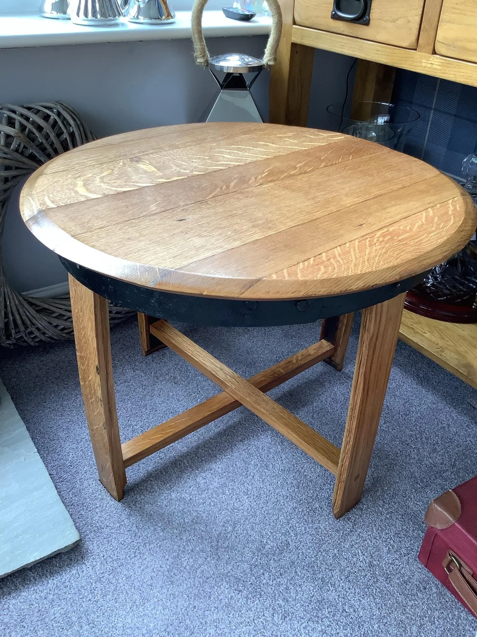 Round wooden table with a black metal rim, supported by four wooden legs and crossbars, situated on gray carpet in a room with wooden furniture and decorative items.