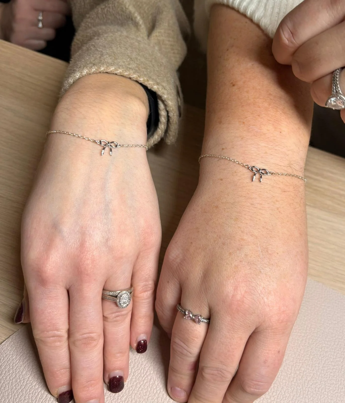 Two hands display matching delicate chain bracelets with small bow charms. Both wear rings; the left hand has a large engagement ring, and the right hand has a smaller ring. One person has dark red nail polish. The hands rest on a light-colored surfa