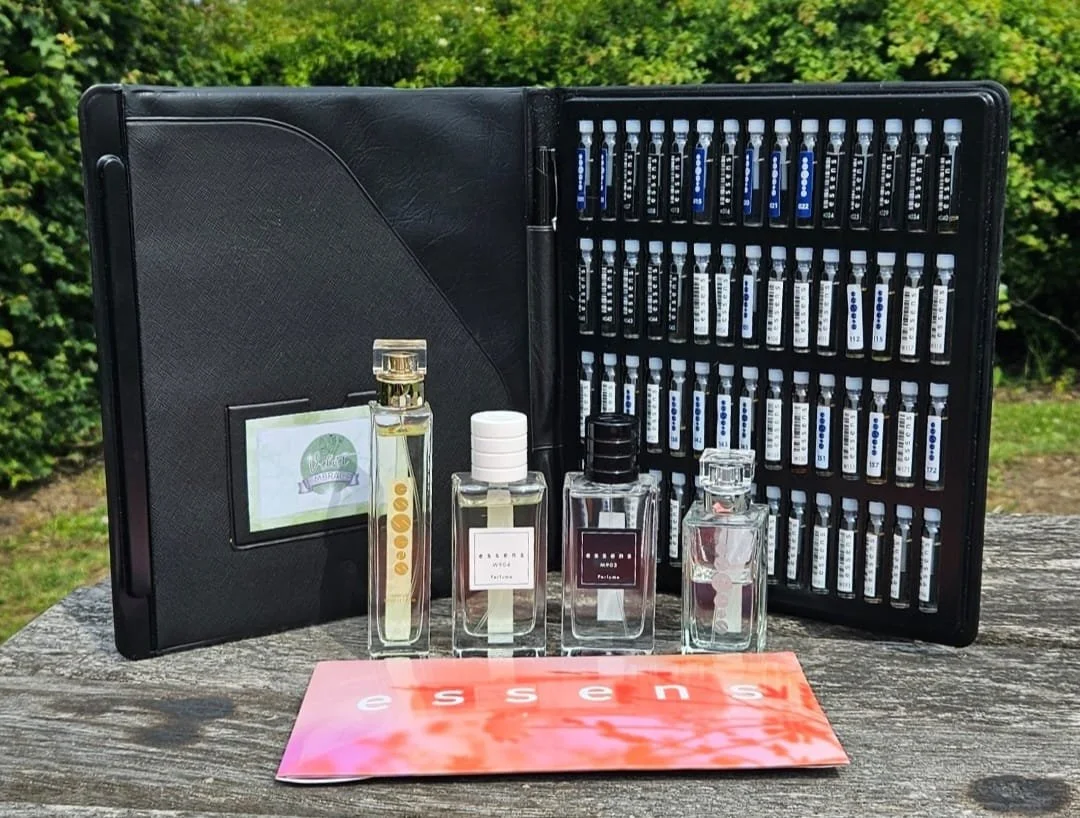 A black scent testing kit case containing numerous small vials of fragrance samples and four larger bottles of perfume outside on a wooden surface with greenery in the background.