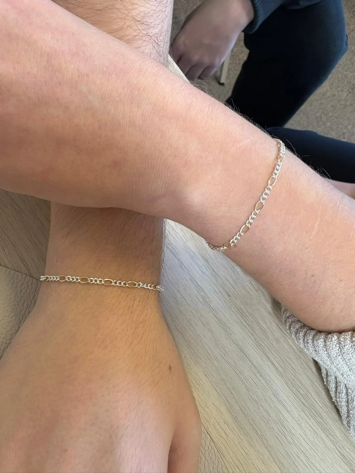 Close-up of two arms, each wearing a silver chain bracelet, resting on a wood table.